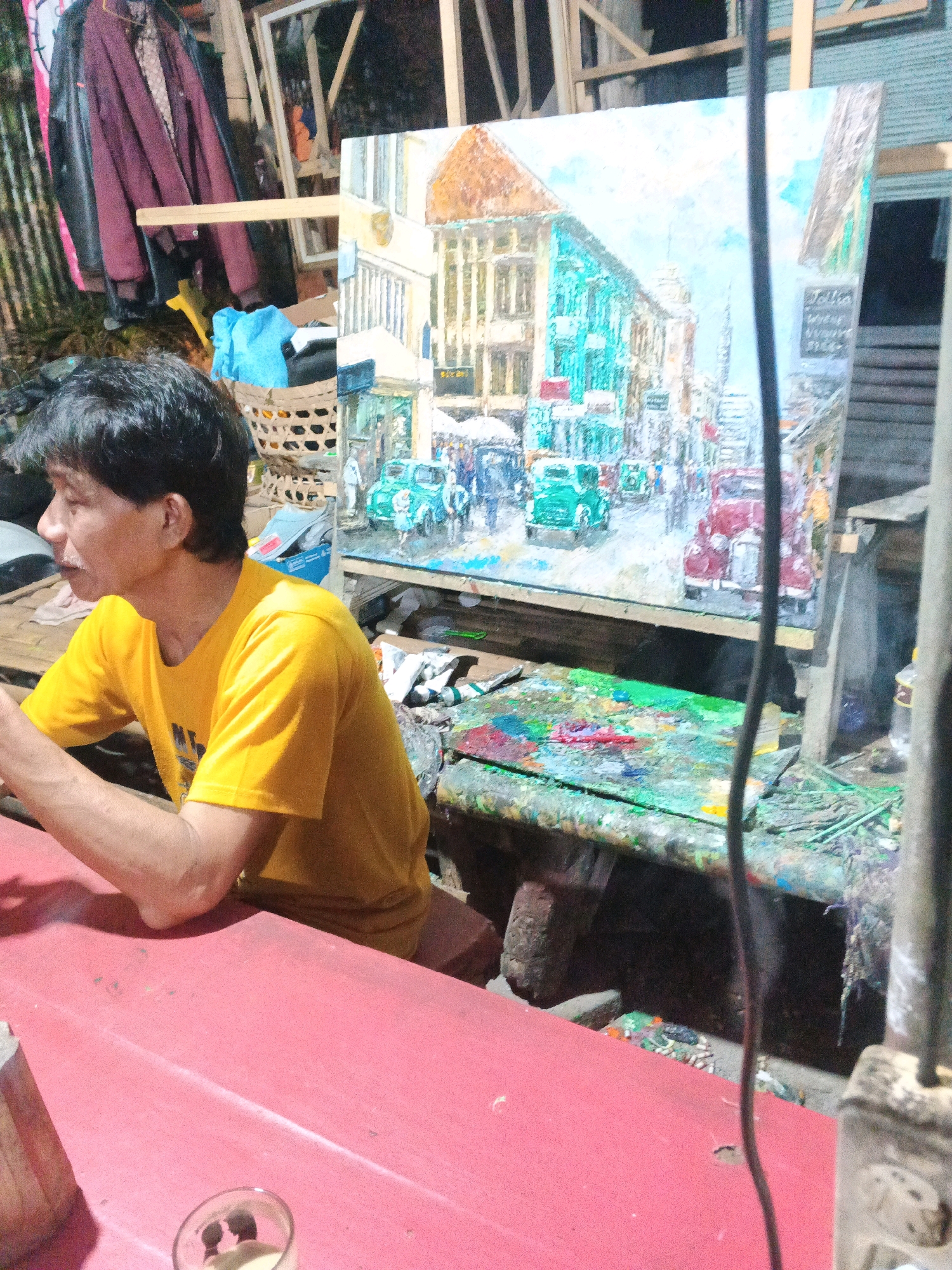 A man in a yellow t-shirt sits at a red table, looking towards a large, colorful painting on an easel. The painting depicts a street scene with buildings and vehicles, likely in a vintage style, and is signed "Jollin". To the left of the man, coats are hanging on a rack, and a woven basket contains a blue cloth. The environment appears to be an outdoor workspace or studio, with paint splatters visible on a workbench in the background. The scene is dimly lit, suggesting it may be evening. The specific location in Ungaran, Indonesia, is not explicitly indicated within the image itself.