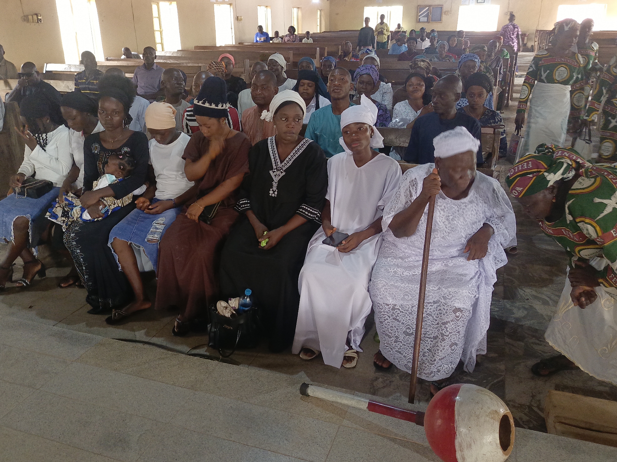 This image captures a large gathering of people, predominantly adults, seated in rows of wooden pews inside what appears to be a church or community hall. Bright light streams in from high windows, suggesting it is daytime.
The subjects include a diverse group of men and women, with some children and a baby visible in the foreground. Many individuals wear head coverings, with some women dressed in white garments, while others wear a mix of traditional Nigerian attire and contemporary casual clothing, such as jeans. One woman in the front row cradles a baby. An elderly woman, also in a white lace garment, holds a wooden walking stick.
The event taking place appears to be a community or religious gathering, possibly a church service or a meeting. People are seated and generally attentive, with many looking towards the front or side, while some glance at the camera.
Notable details include a large, red and white painted gourd-like object, likely a traditional musical instrument, resting on the floor in the foreground. A woman standing in the aisle on the right wears a distinctive green, white, and red patterned dress. Text visible on this dress includes "FELLOWSHIP," "WOMEN," "YEHOVA," "ELIM," and "AKWA IBOM," indicating a group or organization within a religious context. The overall atmosphere is calm and orderly, typical of a communal assembly in Akwanga, Nigeria.