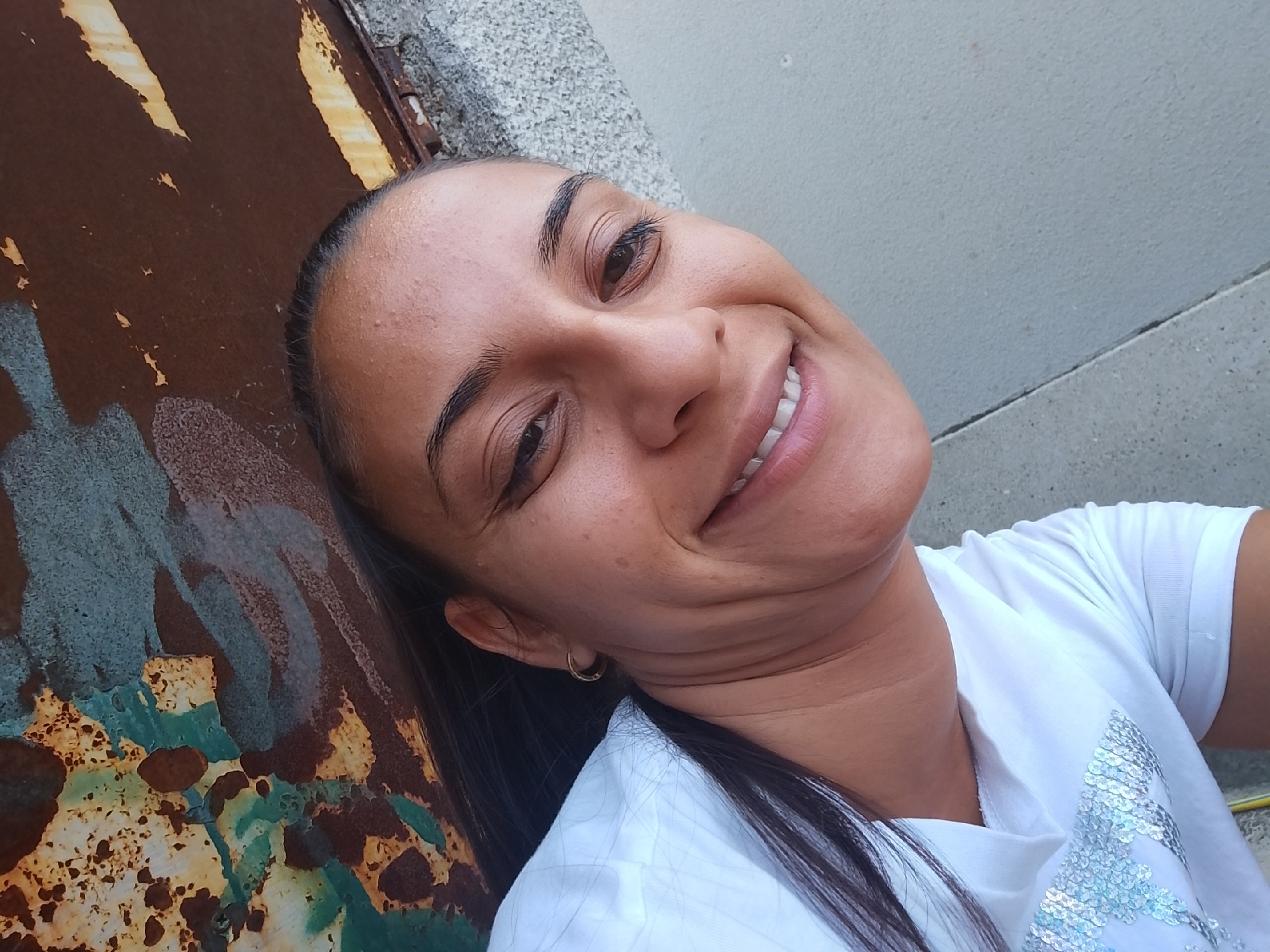 A woman is taking a selfie outdoors, with her head tilted back and her face turned towards the camera. She is smiling broadly, revealing her teeth. Her dark hair is pulled back, and she is wearing a white t-shirt adorned with silver sequins. She has a small hoop earring in her visible ear.
The background of the image is a textured wall with some graffiti and a rusty, weathered metal surface. The lighting suggests it is daytime, possibly in the afternoon, with shadows indicating the sun is at an angle. The overall impression is a casual, candid moment captured in an urban environment, potentially in Yambol, Bulgaria, given the provided context.