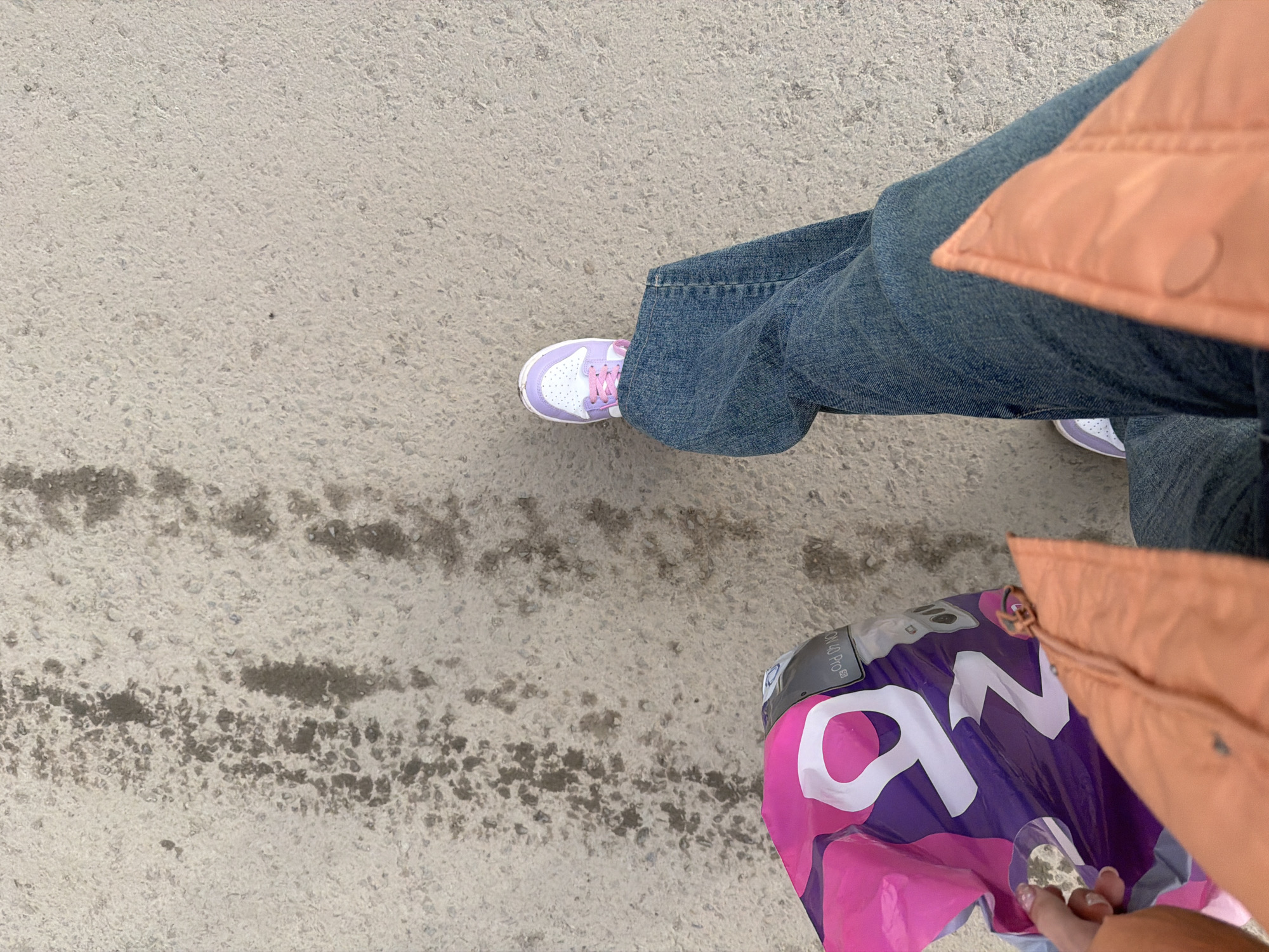 The image shows a person standing on a gray concrete surface, possibly a sidewalk or street in the city of Berezniki, Russia. The person is wearing blue jeans and a peach-colored jacket. They are wearing white and lavender sneakers. They are holding a purple and pink plastic shopping bag with white lettering on it. The bag features "ON LAGO PRO" logo. The concrete surface appears slightly damp in one area. The angle of the shot is from above, looking down.