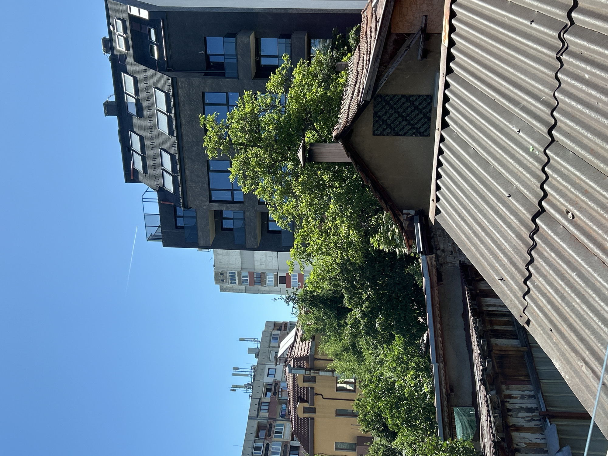 The image shows a view of Sofia, Bulgaria, featuring a mix of old and modern architecture. In the foreground, there is an old building with a corrugated metal roof and dilapidated wooden structures. Adjacent to the old building is a more modern gray building with multiple floors and rectangular windows. Green trees are interspersed between the buildings. In the background, other residential buildings of varying heights and colors are visible under a clear blue sky. A contrail from an airplane can be seen in the sky.