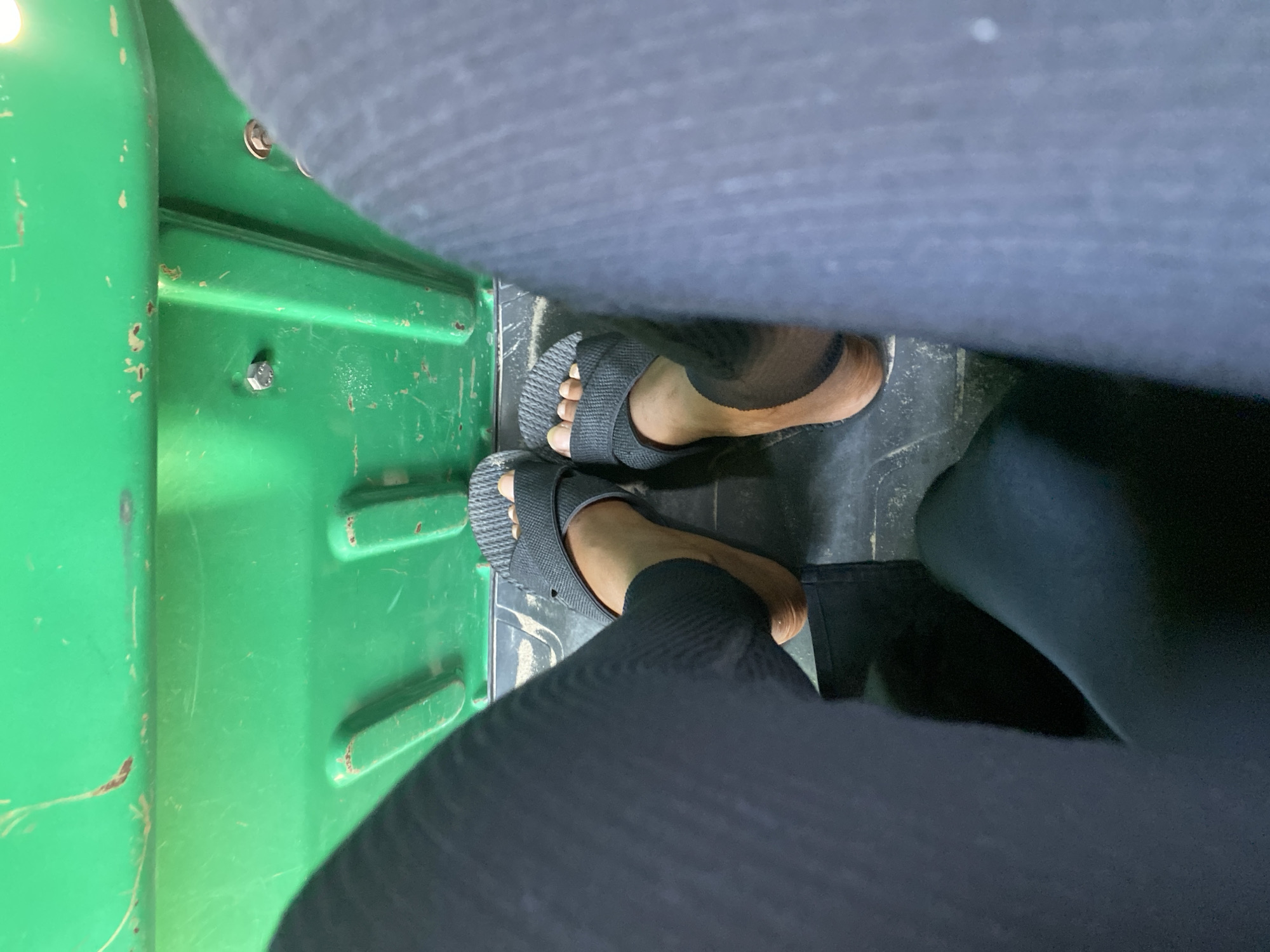 The image shows a person's feet wearing gray sandals inside a vehicle with a green interior. The person is wearing black pants. The floor of the vehicle is black. The green interior has visible wear and tear. There is no information available to identify the location in Dar es-Salaam, Tanzania.