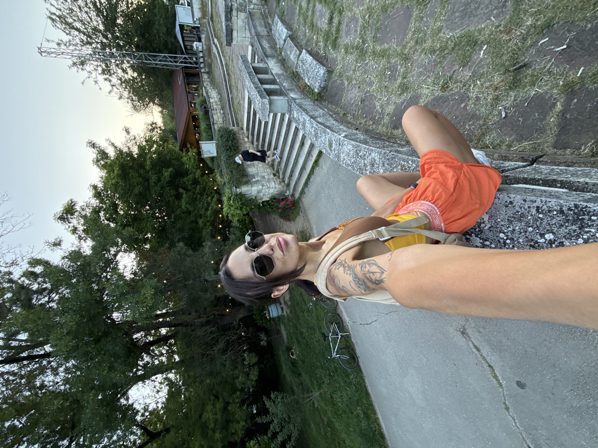 In the city of Pleven, Bulgaria, a woman is captured in a selfie, seated on a stone wall. She is wearing sunglasses, a brown tank top, and bright orange shorts. A backpack is slung over her shoulder, and tattoos are visible on her arm. Her legs are bent, and she is looking towards the camera. Behind her, a pathway leads to stone stairs, with a building with a red roof visible in the distance. A person in a black shirt and blue shorts is walking up the stairs. Two bicycles are parked on the grass to the left. The environment appears to be an outdoor park or recreational area, with lush green trees and clear skies. The lighting suggests it is either early morning or late afternoon.