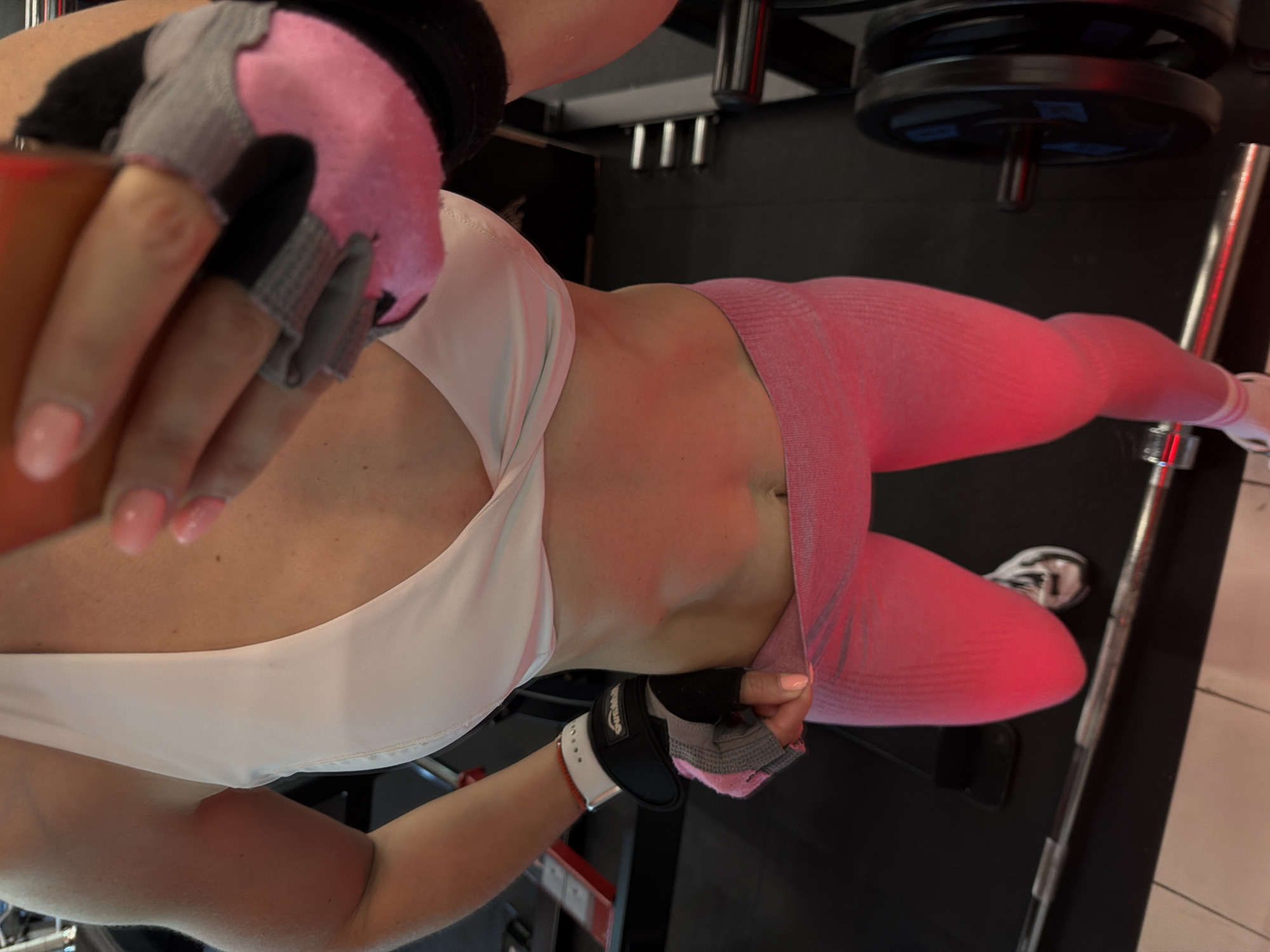 The image shows a person in a gym wearing a white sports bra and pink leggings. They are holding a phone and wearing workout gloves. In the background, there are weightlifting machines and weights. The gym is dimly lit. The scene is a workout session in a gym.