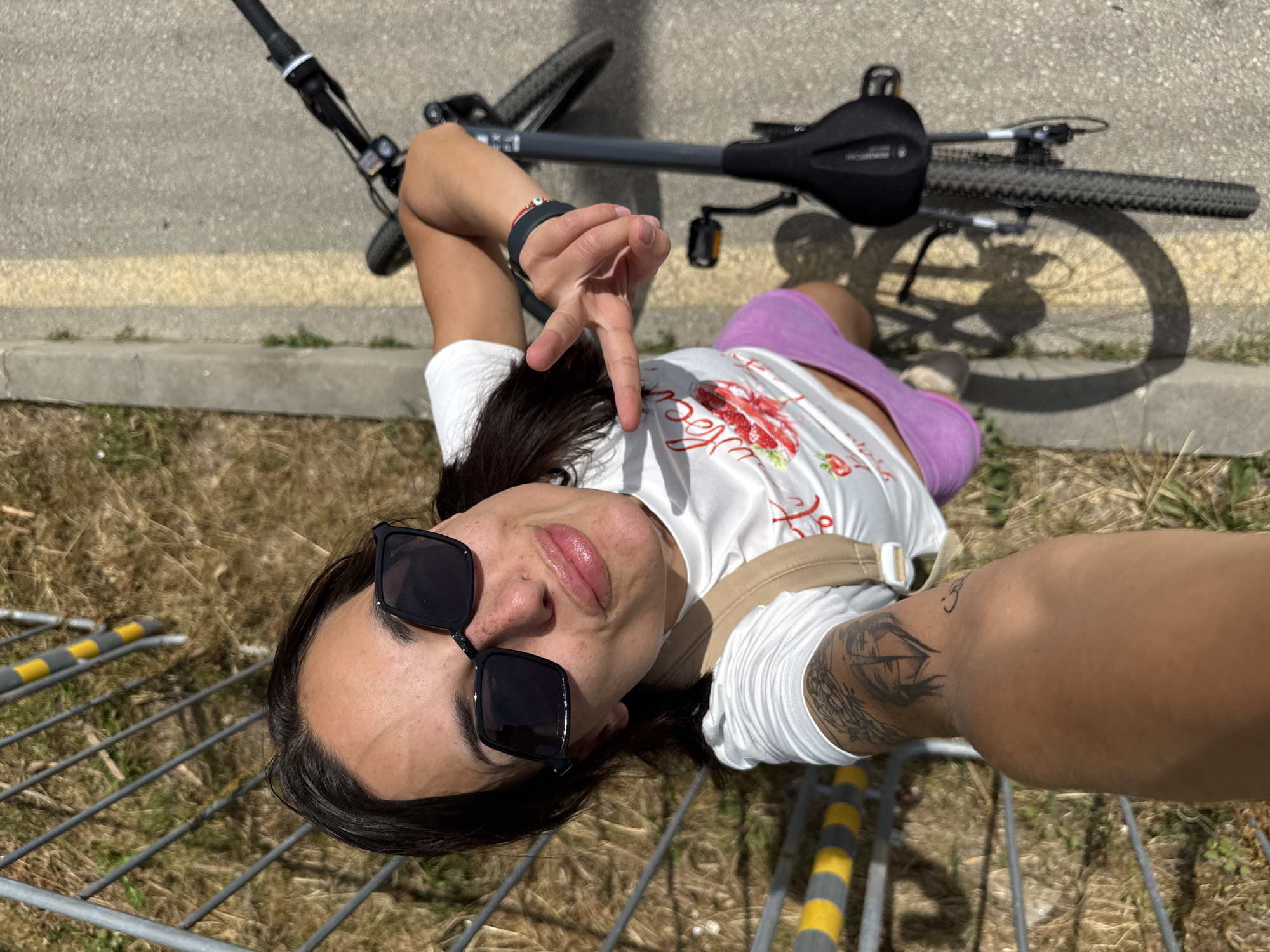 The media file is a selfie taken by a person lying on their back, with a bicycle in the background. The person is wearing a white t-shirt with a floral print, purple shorts, and sunglasses. They are making a peace sign with their right hand. A tattoo is visible on their left arm. The scene appears to be outdoors, with a paved surface and some dry grass. A metal barrier with yellow and grey markings is also visible in the foreground. The lighting suggests it is daytime, likely sunny. The person's expression is relaxed and playful. The bicycle is positioned with its wheels on the ground. The overall impression is of a casual, outdoor activity.