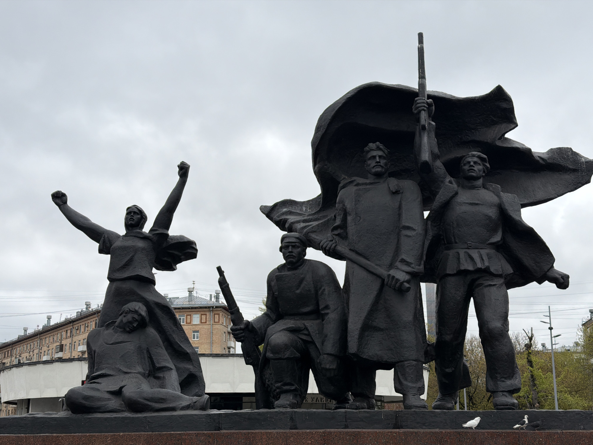 The image depicts a bronze monument with several figures in Moscow, Russia. A woman stands with raised arms, and a fallen figure is seated near her. Three male figures stand with weapons, one holding a rifle overhead. A large, dark fabric appears behind them. The monument sits atop a stone base. In the background is a building and a cloudy sky. Two pigeons are present at the base of the monument.