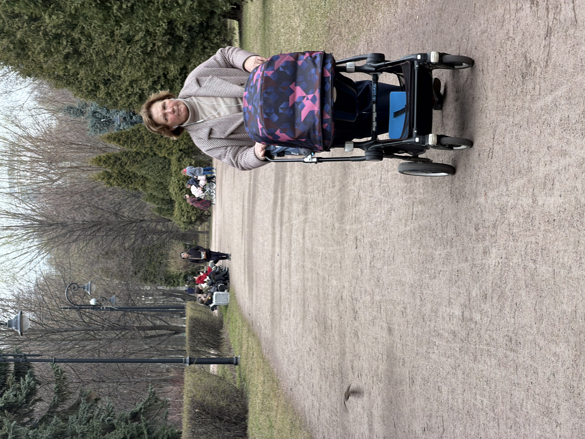An older woman wearing a beige turtleneck and a grey and white plaid jacket pushes a black stroller with a geometric patterned cover. The stroller appears to have a blue container in it. She is on a dirt path in a park with benches on the left where people are sitting. There are bare trees and evergreens on the left of the path and green shrubs. In the background are more people. The path is in a park in Saint Petersburg, Russia.