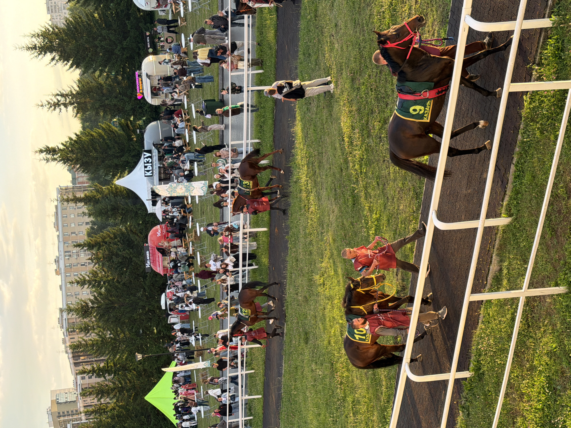 An eye-level, long shot captures a sunny outdoor event with a crowd of people, food stalls, and horses on a race track. The foreground features a white railing separating a grassy area from a dark race track, where two horses with riders are walking. The horse on the right wears a green saddle blanket with the number '9' and the word 'КАЗАНЬ' (KAZAN) in yellow. The horse on the left has a similar saddle blanket with the number '10'. In the midground, a group of people are gathered around food stalls and tents, with trees and apartment buildings in the background. The sky is bright and hazy, suggesting it is late afternoon.