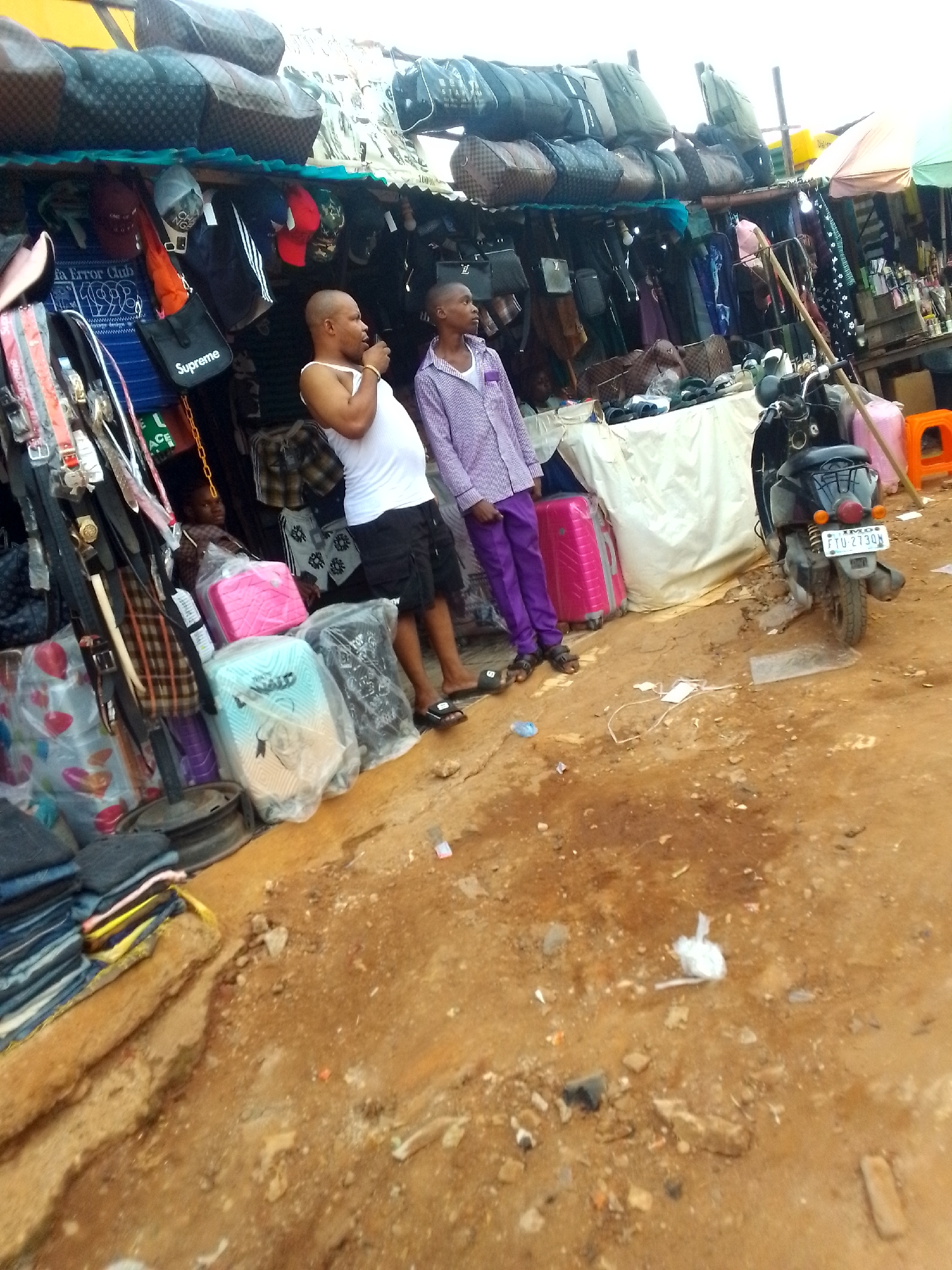 The image depicts a street scene in Orlu, Nigeria. Two men are standing outside a shop selling bags, clothing, and accessories. One man is wearing a white tank top and black shorts, and the other is wearing a purple patterned shirt and purple pants. Suitcases in pink, blue and grey are also displayed for sale. A motorcycle is parked nearby, with the license plate visible "IMO FTU-2730A". The ground is dirt and strewn with litter. Merchandise hangs overhead from the shop's awning, including handbags and clothing.