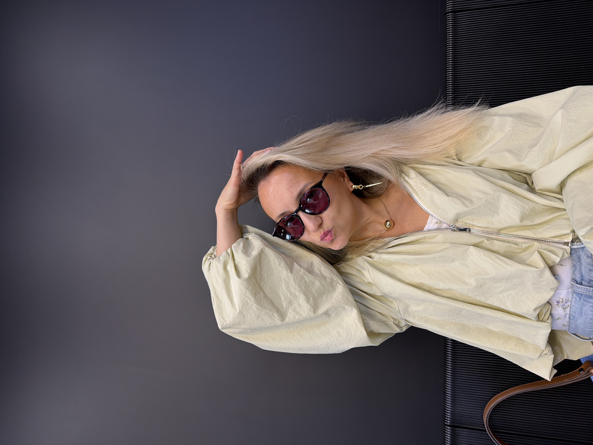 A young Caucasian woman with blonde hair and sunglasses is sitting in front of a dark gray wall, with a ribbed black bench in the foreground. The woman is wearing a light yellow hooded jacket with voluminous sleeves, a white top, and light blue jeans. She has her right hand on her head and is sticking out her tongue. She is wearing a gold spherical pendant necklace and a dangling earring. The scene appears to be indoors, possibly in a cafe or a waiting area. There is no indication that the location is in Izhevsk, Russia.