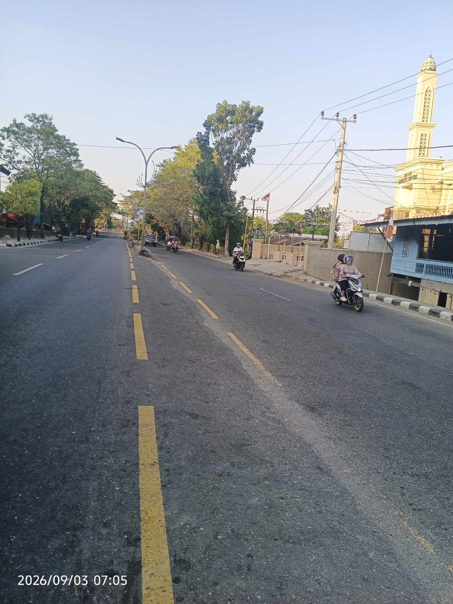 The image captures a street scene in Pare-Pare, Indonesia, with a focus on a road lined with trees and buildings. A light blue building and a tall, cream-colored minaret are visible in the background on the right, suggesting the presence of a mosque. Several people are riding motorcycles on the road. One motorcycle in the foreground carries two individuals, a driver and a passenger, both wearing helmets. Another motorcycle is further down the road to the left, with a single rider. The road is paved and has yellow lane markings. Overhead power lines and streetlights are also present. The sky is clear and appears to be a bright, sunny day, possibly late afternoon or early morning given the lighting. The date stamp at the bottom left corner indicates September 3, 2026, at 07:05.