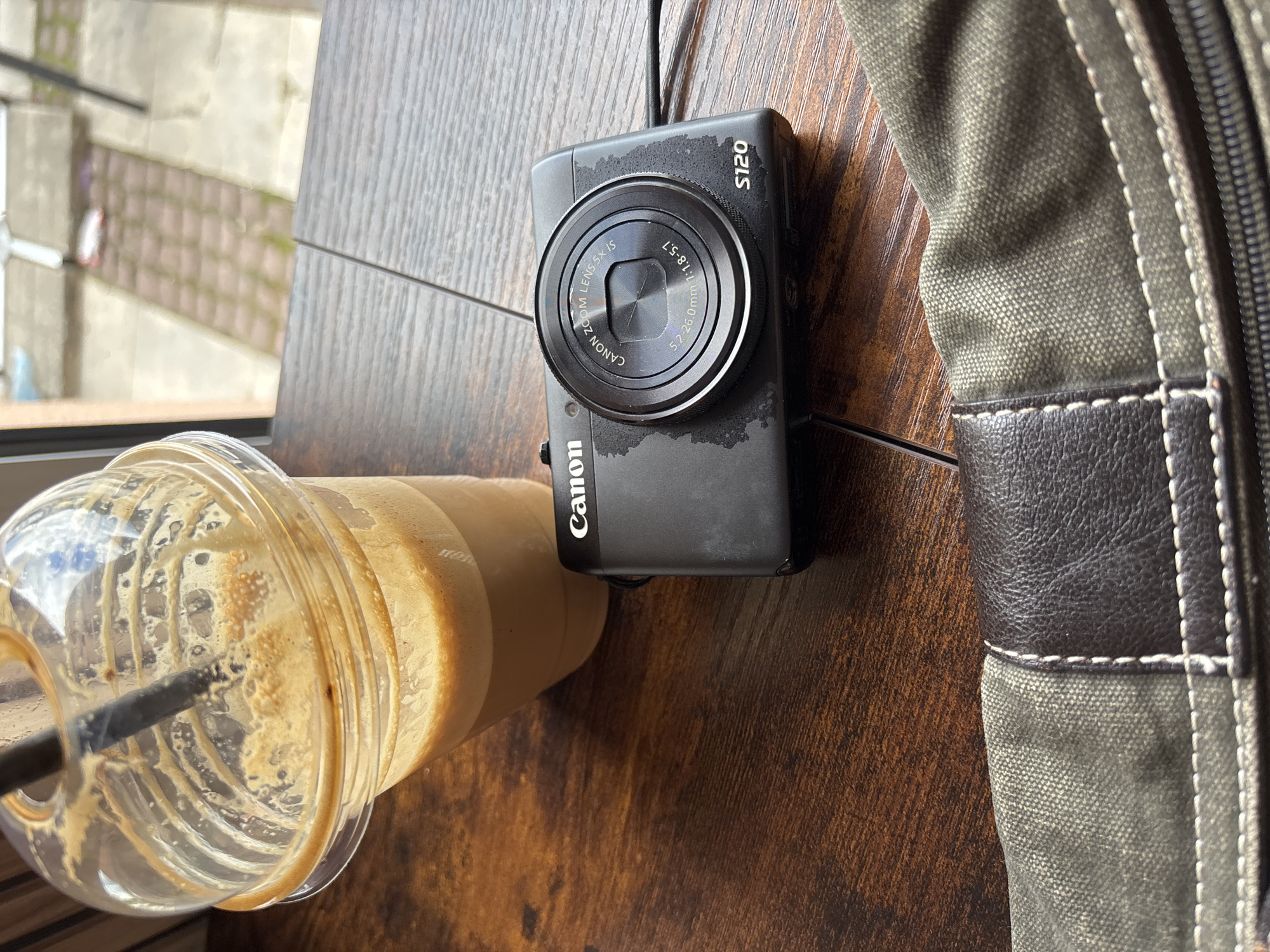 The image shows a table in Garmen, Bulgaria with a half-empty plastic cup of iced coffee, a Canon S120 digital camera, and a canvas bag. The table is made of dark brown wood. A window with a view of the street and a building is visible. No people are visible. The coffee cup has a black straw in it. The bag is olive green with a brown leather trim and a zipper. The camera is black and shows signs of use, with visible smudges.
