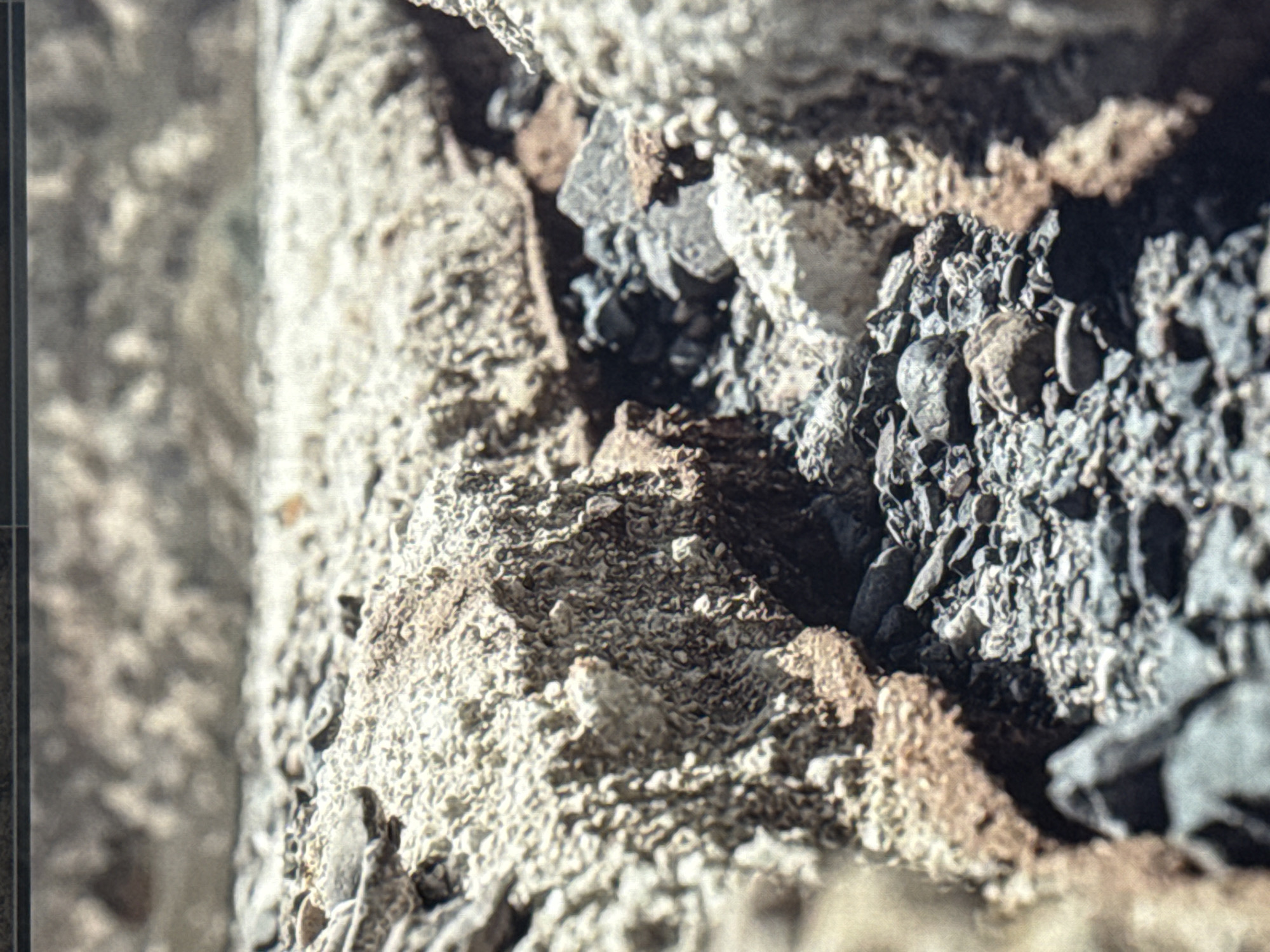 This is a close-up, macro shot of a rough, textured surface composed of small stones, gravel, and what appears to be concrete or dried mud. The color palette is dominated by earthy tones of gray, beige, and hints of brown, with some darker, almost black pebbles mixed in. The texture is highly varied, with some areas appearing porous and granular, while others are comprised of sharp, angular fragments.
The lighting suggests it might be daytime, with shadows indicating a light source from the upper right. The focus is shallow, with parts of the image being sharp and detailed, while other areas are blurred, creating a sense of depth and emphasizing the tactile nature of the material. The composition is abstract, focusing on the geological or constructional elements rather than any specific recognizable object or scene. There is no visible text, people, or discernible activity. The overall impression is one of ruggedness and natural or man-made decay, possibly a fragment of a wall, road, or rocky terrain. Without further context, pinpointing the exact location within Eyüpsultan, Turkey, is difficult, but the materials suggest an outdoor, possibly urban or construction-related environment.