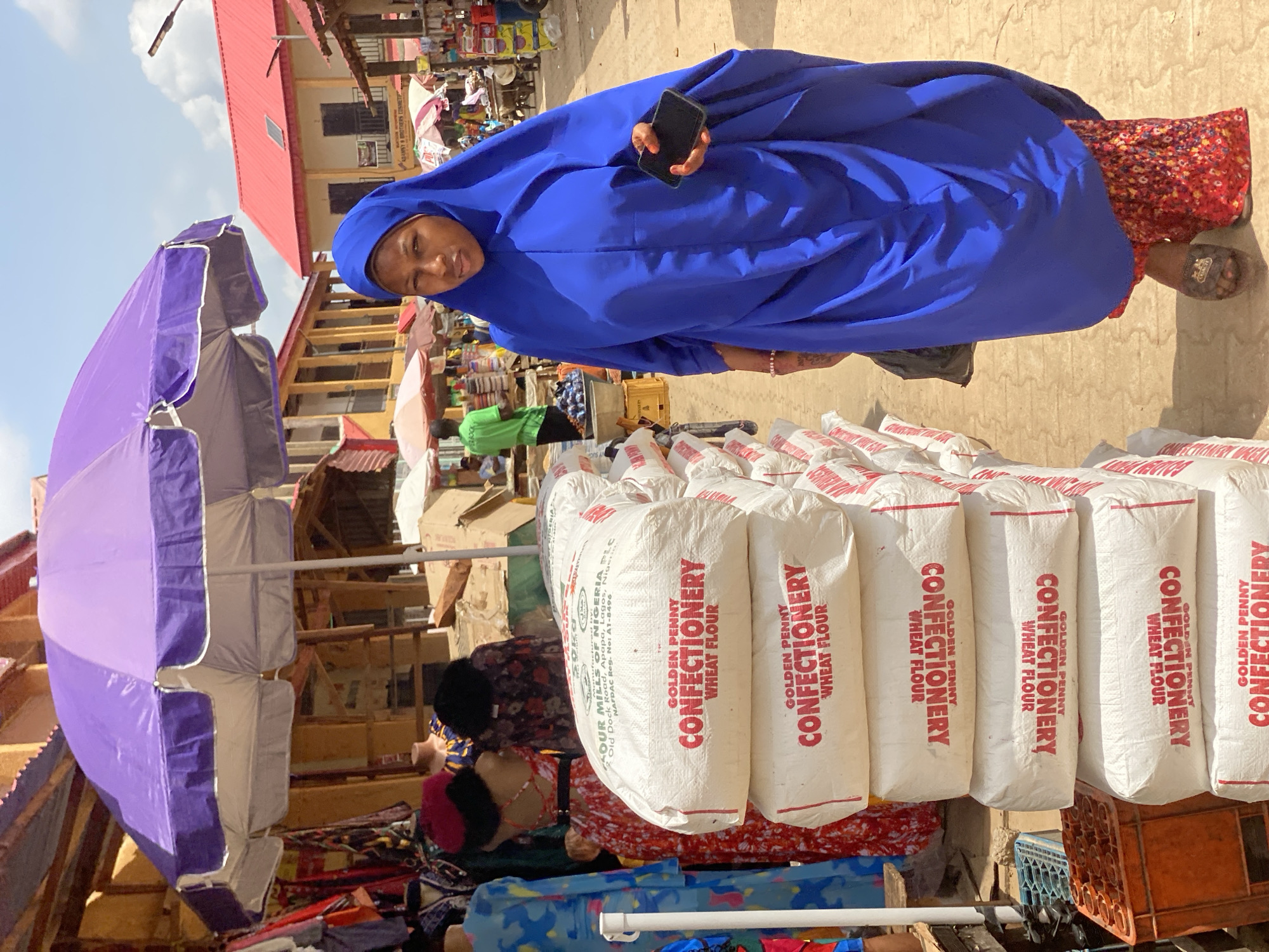 A woman in a blue garment holds a phone in an outdoor market setting. She stands near a stack of white bags labeled "GOLDEN PENNY CONFECTIONERY WHEAT FLOUR." The market features stalls with purple umbrellas, various goods, and other people. The buildings in the background have red roofs and multiple stories. The scene is in Kafanchan B, Nigeria.