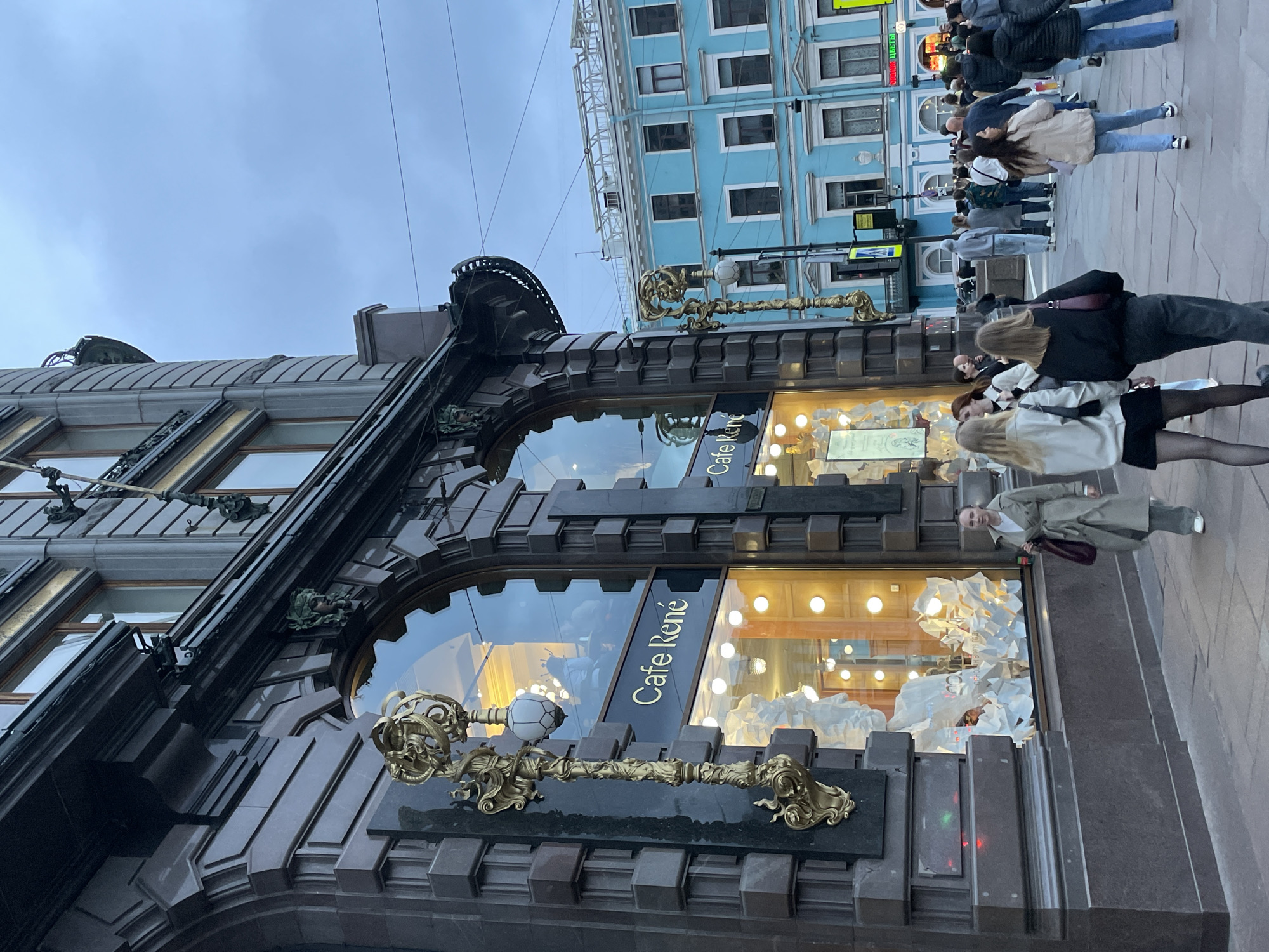 The image depicts a street scene in Saint Petersburg, Russia. The focal point is the storefront of "Cafe René," characterized by its dark, ornate architectural style with large arched windows and decorative golden embellishments. A long, ornamental golden key design is affixed to the building facade. The cafe's windows display a light-colored interior with what appear to be sculptural decorations. A small group of people is visible on the sidewalk in front of the cafe, partially obscured. Behind the cafe, across the street, is a light blue building with rows of windows. The sky is overcast, creating a muted lighting environment.
