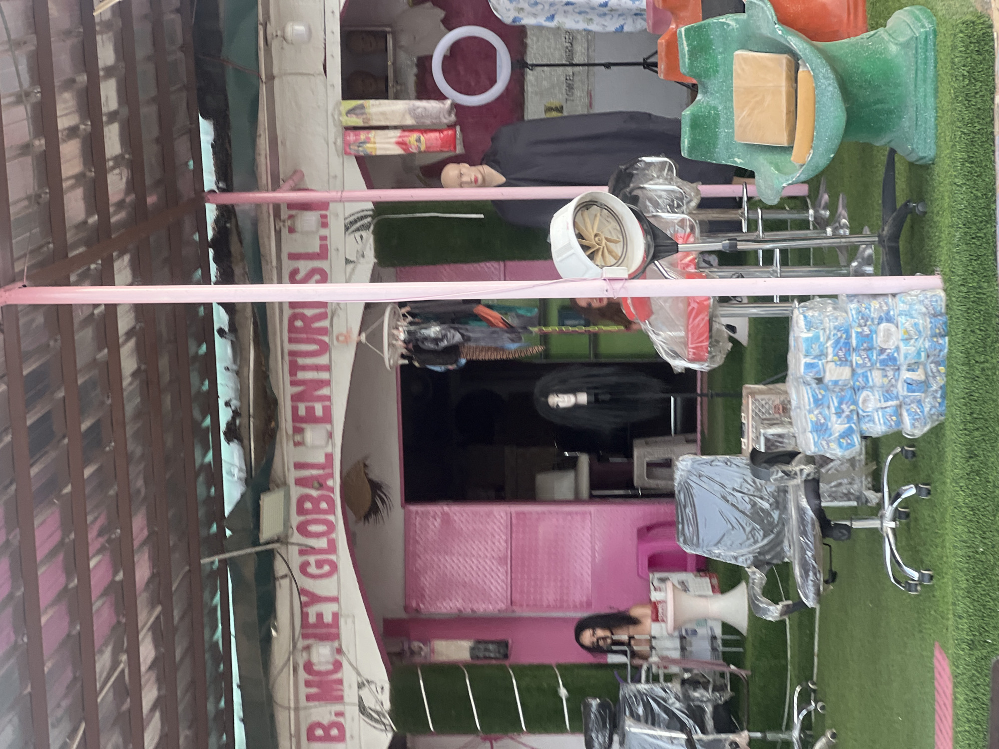 This is a full-frame, eye-level, outdoor, daytime photograph of a storefront in Jalingo, Nigeria, likely a salon or beauty supply shop. The storefront is painted pink and white and features a corrugated metal roof supported by wooden beams. The name "B. MONEY GLOBAL VENTURES LTD." is prominently displayed in pink lettering above the entrance.
In front of the store, several items are displayed on artificial grass. These include office chairs covered in plastic, stacks of packaged items (possibly toiletries or cleaning supplies), a salon-style hair dryer on a stand, and two decorative green seats, one holding a brown package and a yellow cushion.
Mannequins are also present. One mannequin on the left, wearing a white top, is partially visible behind a plastic-covered chair. Another mannequin to the right, wearing a black robe and a red lipstick, stands behind a pink metal support pole. A third mannequin with long, black hair is visible in the doorway of the shop. A ring light on a tripod is positioned to the right of the black-robed mannequin.
The background shows the interior of the shop, which is dark and appears to contain more salon equipment. The overall scene suggests a commercial establishment preparing for or operating during business hours.