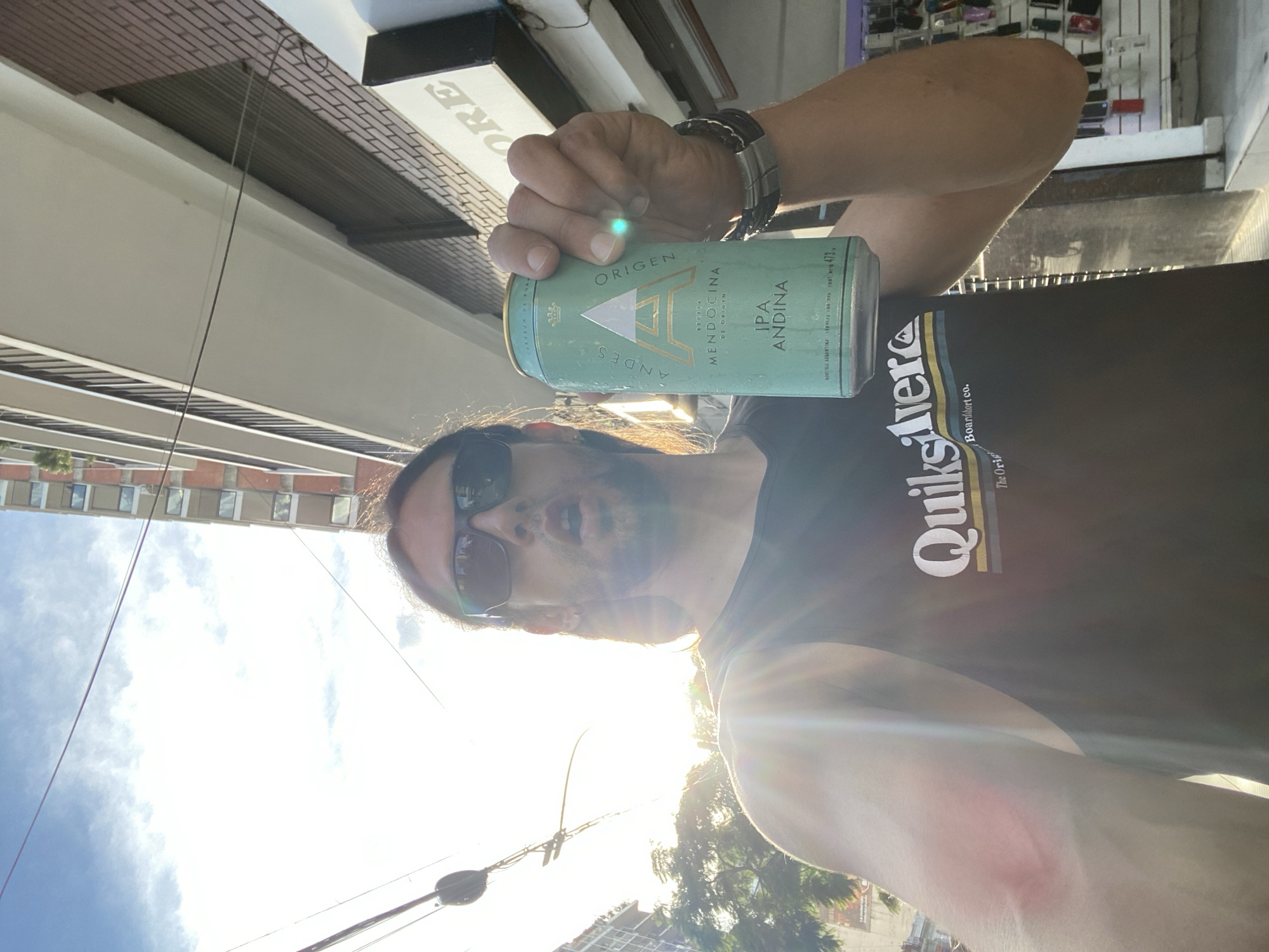 A man with long hair and sunglasses is holding a can of "Origen Andes IPA Andina" beer. He is wearing a black Quiksilver t-shirt and a watch. The background includes buildings, power lines, a storefront, and the sky. The location appears to be an urban area, possibly in Buenos Aires, Argentina. The photo is taken during a sunny day with the sun in the background.