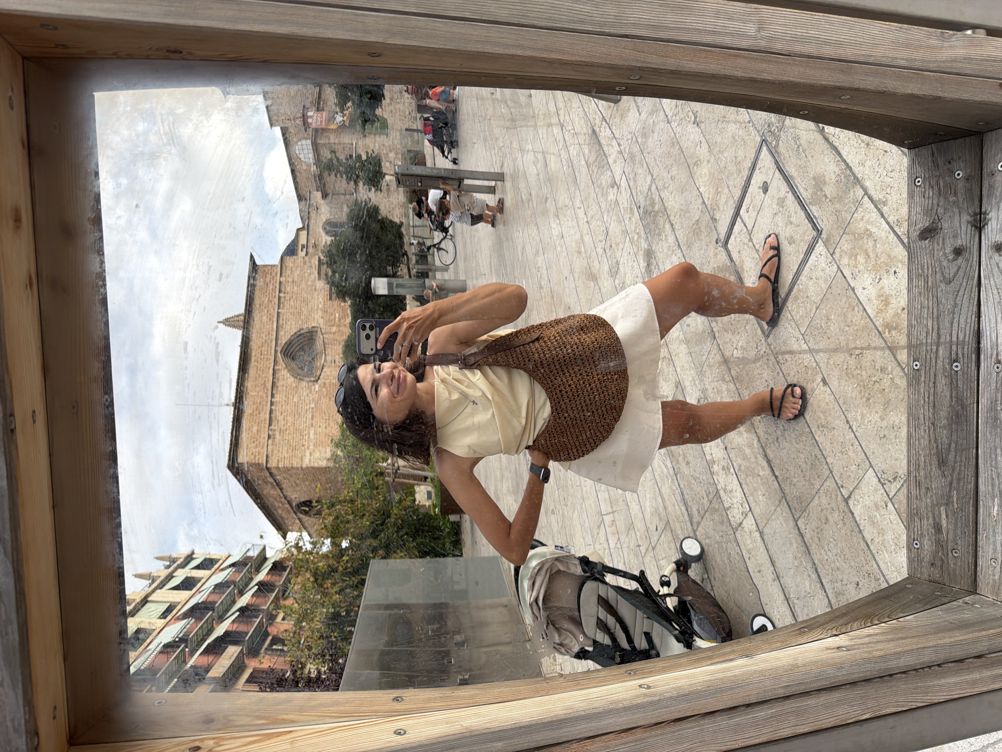 A woman with short brown hair takes a mirror selfie in a tiled urban plaza. She is wearing a cream-colored sleeveless top with a draped neckline, a white mini skirt, and black strappy sandals. A brown woven bag hangs over her right shoulder. Her left hand is on her hip, and her right hand holds a smartphone. In the reflection, a baby stroller is visible to her left. In the background, there are brick buildings, trees, and other people, including a child interacting with a pole and another child in a stroller. The plaza has a light-colored tiled floor with a square metal grate. The sky is overcast. The mirror is framed by rough wooden planks.