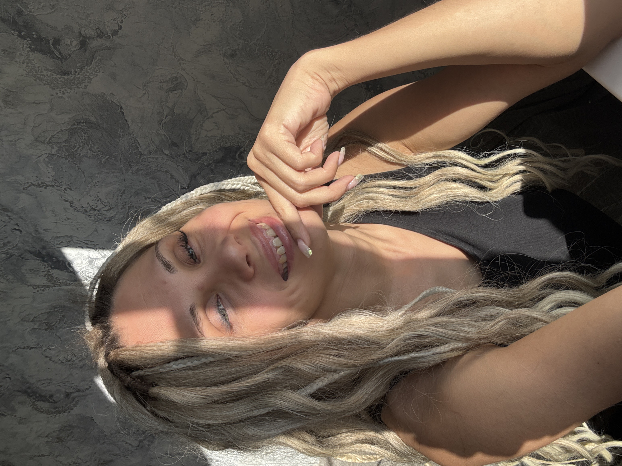 A fair-skinned woman with blonde, braided hair is smiling at the camera. She has her right hand near her face, with her fingers curled inwards. She is wearing a black top. The background appears to be a grey, textured surface. There is a strong light source creating sharp shadows across her face and the background. The location is not discernible from the image.