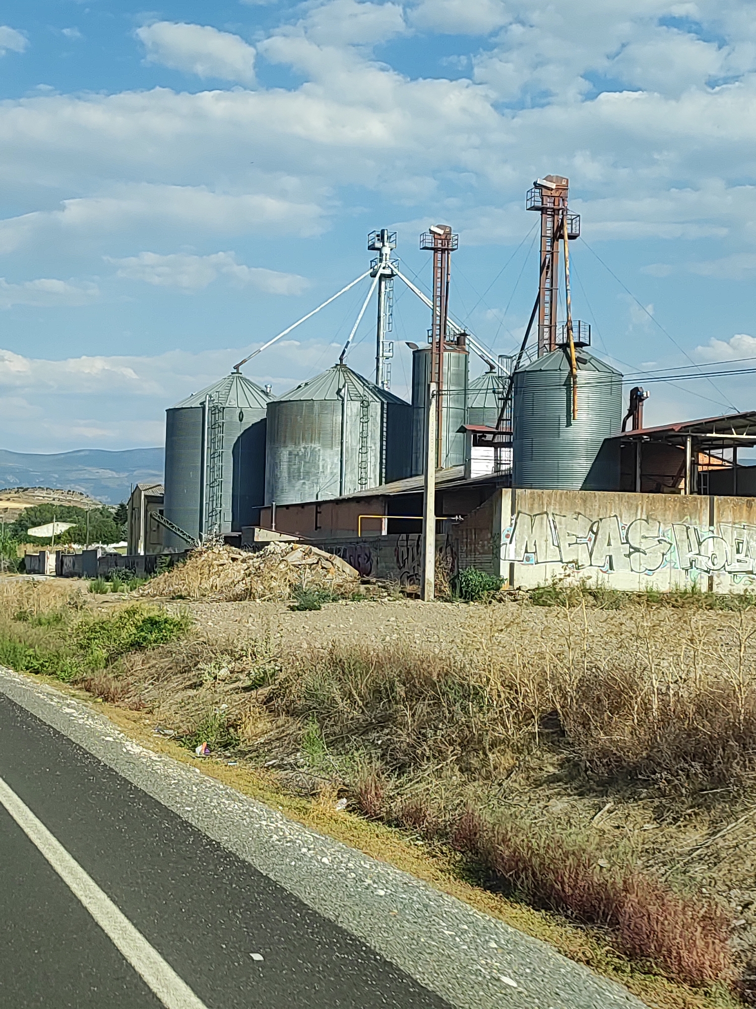 An industrial scene unfolds under a partly cloudy blue sky. The primary focus is a row of tall, cylindrical grain silos, some appearing weathered and metallic, others a dull gray. Several of these silos are connected by a network of white metal bracing and chutes. Atop some silos are tall, rusted metal structures with platforms and what appear to be loading mechanisms.
To the right of the silos, a low-slung building with a corrugated metal roof is visible. A concrete wall, covered in graffiti that includes the word "MEASURE," runs along the foreground. The ground leading up to the silos is uneven and covered with dry grass, dirt, and scattered debris. A steep embankment rises behind the silos, dotted with sparse vegetation and trees, with rolling hills in the distance.
In the immediate foreground, a section of asphalt road with a white lane marker is visible, suggesting the scene is located alongside a road. The overall impression is one of an agricultural or industrial facility in a somewhat neglected state. The specific location in Segovia, Spain, is not discernible from the visual cues within the image itself.