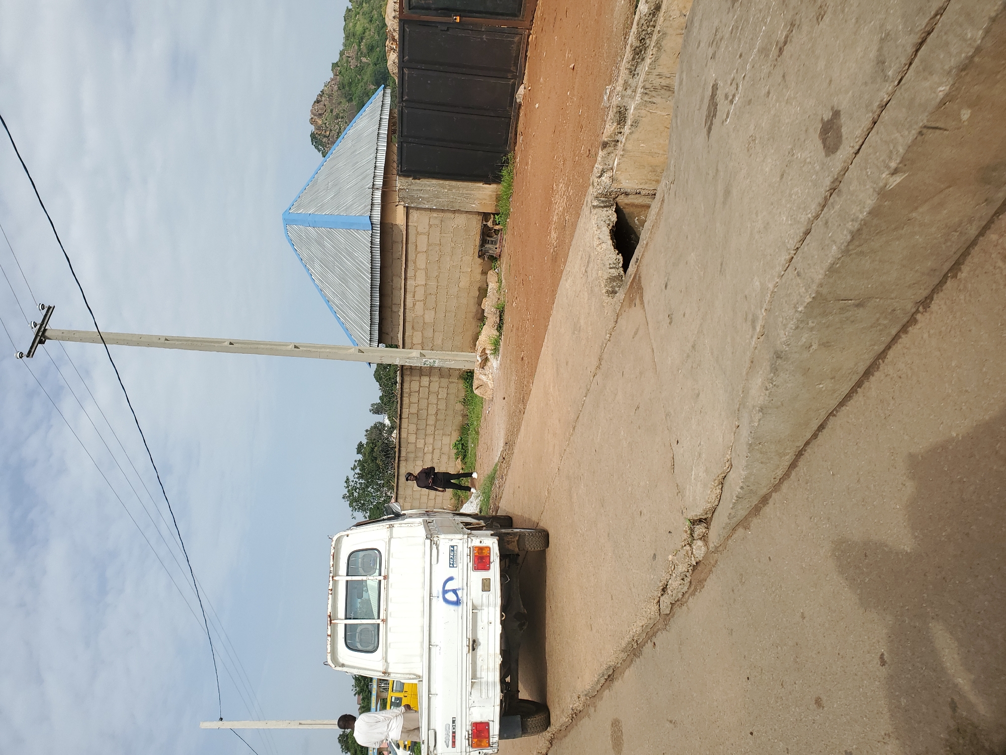 In Bukuru, Nigeria, a white pickup truck is parked on a concrete road. The truck's bed is empty. Two men are visible: one stands to the left of the truck, facing away from the camera and wearing a light-colored shirt and shorts. The second man stands to the right of the truck, facing towards the truck and wearing dark clothing. In the background, a brick wall runs along a dirt path, and a large, dark metal gate is visible. A building with a silver, corrugated metal roof trimmed in blue sits behind the wall. A concrete utility pole with electrical wires extends upwards into a cloudy sky. The road has a visible concrete curb and a small drainage ditch.