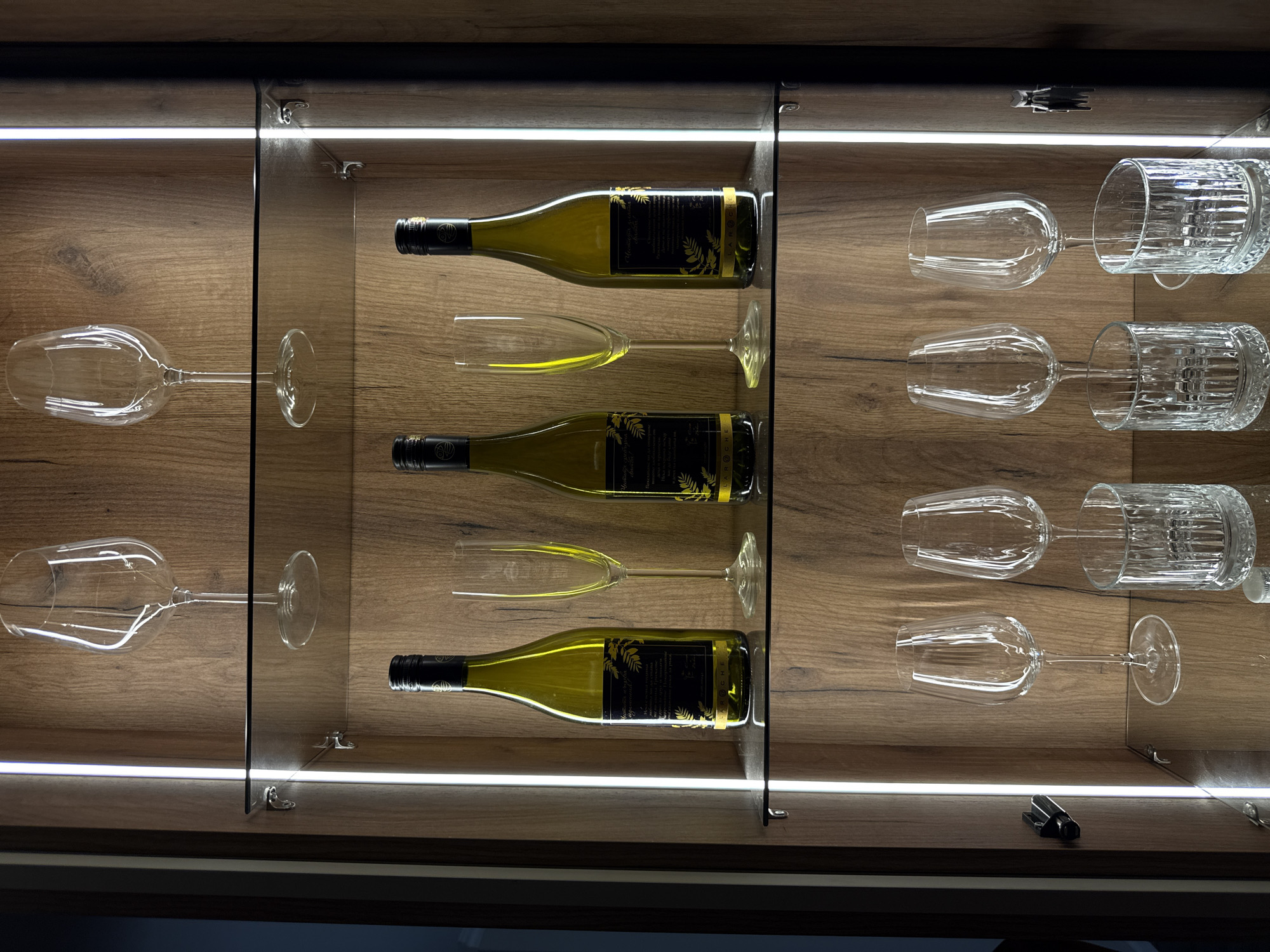 The image displays a well-lit wooden cabinet with glass shelves, showcasing an assortment of wine bottles and glasses. The cabinet is illuminated by vertical LED strip lights on either side. On the top shelf, two tall, clear wine glasses are placed symmetrically. The middle shelf holds two green wine bottles, each accompanied by a tall, slender champagne flute. The bottom shelf features four glasses: two goblet-style wine glasses and two short, wide tumblers with ribbed patterns. The cabinet's wooden interior has a prominent grain pattern. There are no people or animals present in the image, and no specific location in Plovdiv, Bulgaria, is discernible from the scene.