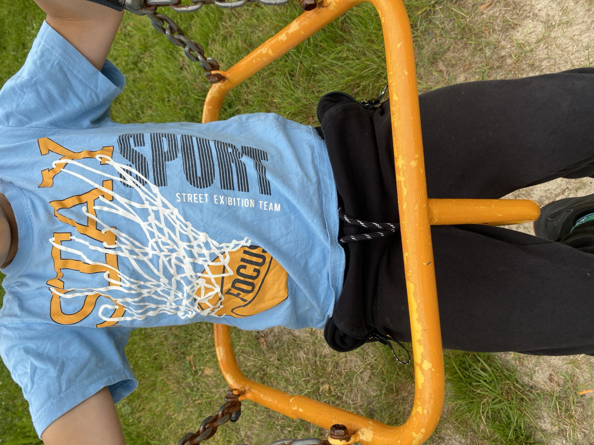 The image shows a close-up, overhead view of a young person sitting on an orange metal swing. The person is wearing a light blue t-shirt with a graphic design featuring the word "SPORT" in bold black letters, with thin white lines resembling a basketball net overlaid. Below "SPORT," the text "STREET EXIBITION TEAM" is visible. To the right of the text, a circular graphic with the word "FOCUS" is partially visible. The person is also wearing black pants. The swing is made of thick, bright orange metal tubing, with rusty chains attached to its frame. The ground below is covered with green grass and some sandy patches. The lighting suggests it is daytime, likely sunny, with no strong shadows. The overall impression is one of a casual outdoor activity, possibly at a playground or park in Chulis, Russia.