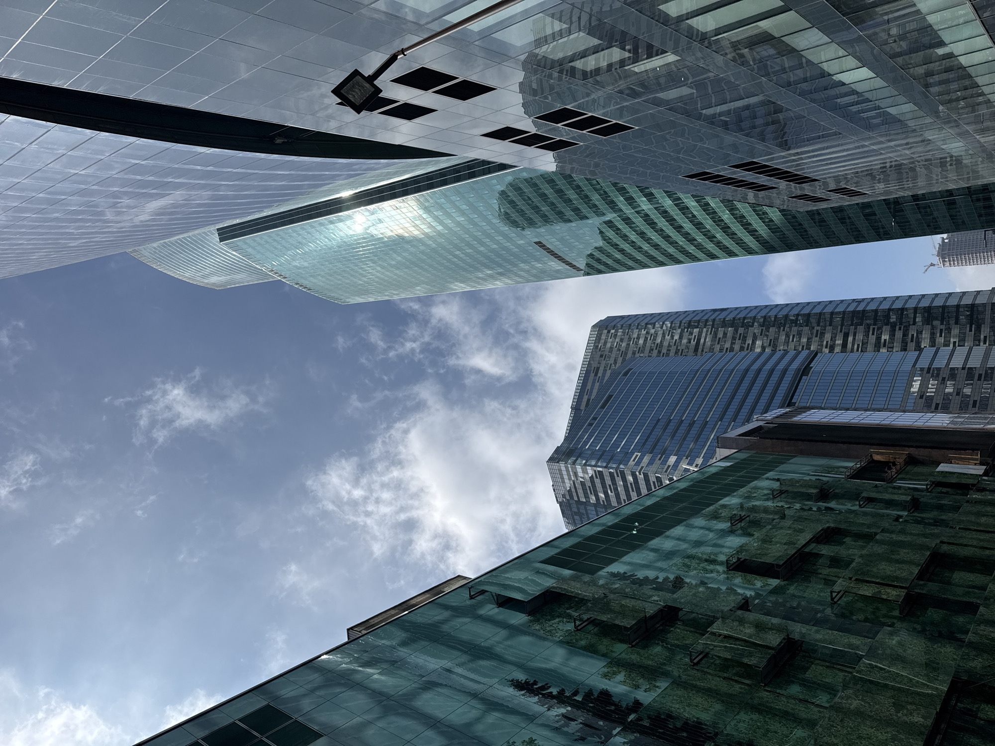 The image shows a view looking up at several tall buildings with glass facades. The buildings are arranged in a cluster, obscuring much of the sky. The sky is visible in the gaps between the buildings and is a light blue color with some white clouds. The buildings have a modern design with clean lines and reflective surfaces. The bottom building shows reflection of tree branches. It is taken in the city of Unknown, Unknown.