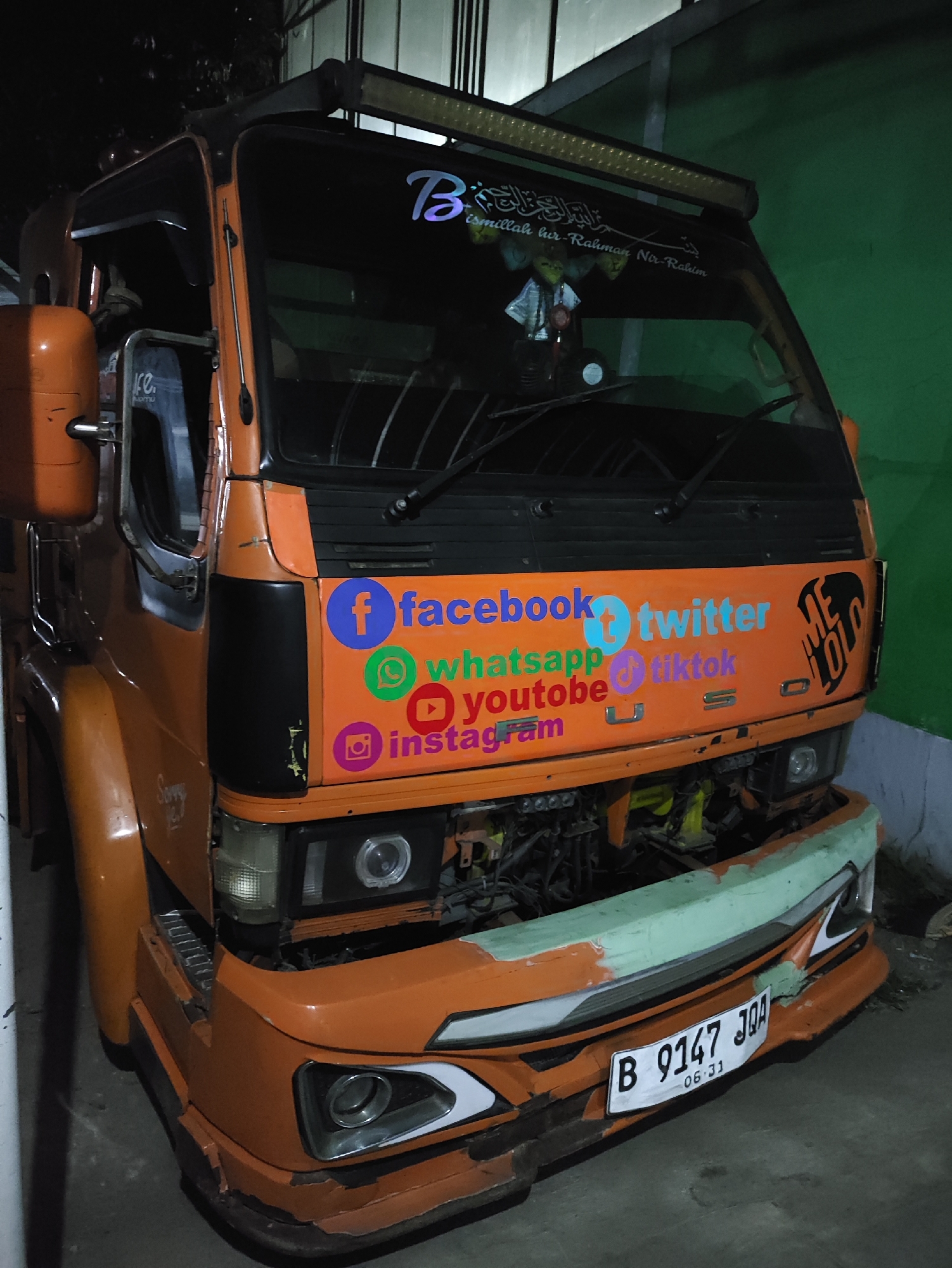 An orange truck is parked in what appears to be a dimly lit garage or alley in Kepung, Indonesia. The truck has several stickers on its front, including the logos for Facebook, Twitter, WhatsApp, YouTube, and Instagram, along with the word "MELOJO" in black lettering. There is also a license plate on the front bumper that reads "B 9147 JQA 06-31". The truck has a light bar on top and is in a state of disrepair, with some parts of the bumper missing and exposed engine components. The background is dark with some visible green wall and ribbed metal structures. No people are visible in the image.