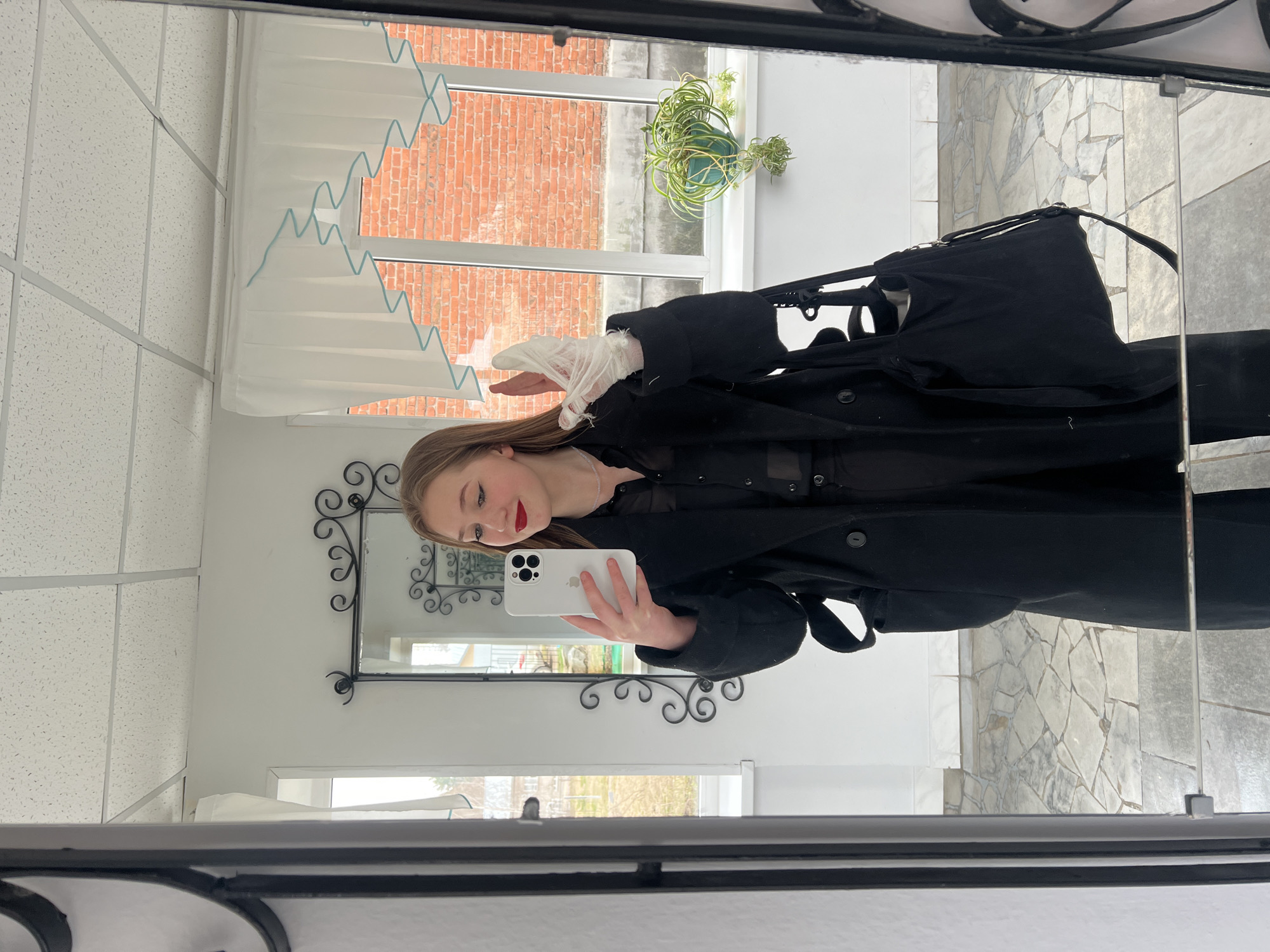 A young woman is captured in a mirror selfie, standing upright and looking towards her white smartphone, which she holds up to take the picture. She has light brown hair, red lipstick, and is wearing a long black coat or trench coat over a sheer black top and pants. A delicate white lace glove or sleeve is visible on her left hand.
The setting, reflected in the large mirror, appears to be an indoor space with various elements. To the left, a portion of a white popcorn-style acoustic ceiling is visible. Behind the woman, a window offers a view of an outdoor brick wall, likely part of an adjacent building, and a green hanging plant in a blue pot. Another smaller window in the reflection shows bare trees and branches outside, suggesting it might be a cooler season.
Inside the room, there are white walls and white pleated blinds adorned with a light blue zigzag pattern. A decorative black-framed mirror with scrollwork is also reflected, situated on the white wall. The floor visible in the reflection is tiled with irregular, light-colored flagstones. The outer frame of the main mirror, encompassing the selfie, is dark, possibly black metal, with some decorative elements at the top. The activity is a casual mirror selfie, likely taken during the daytime given the natural light from the window. No visible text is present. The context provided indicates the location is Chaikovsky, Russia.
