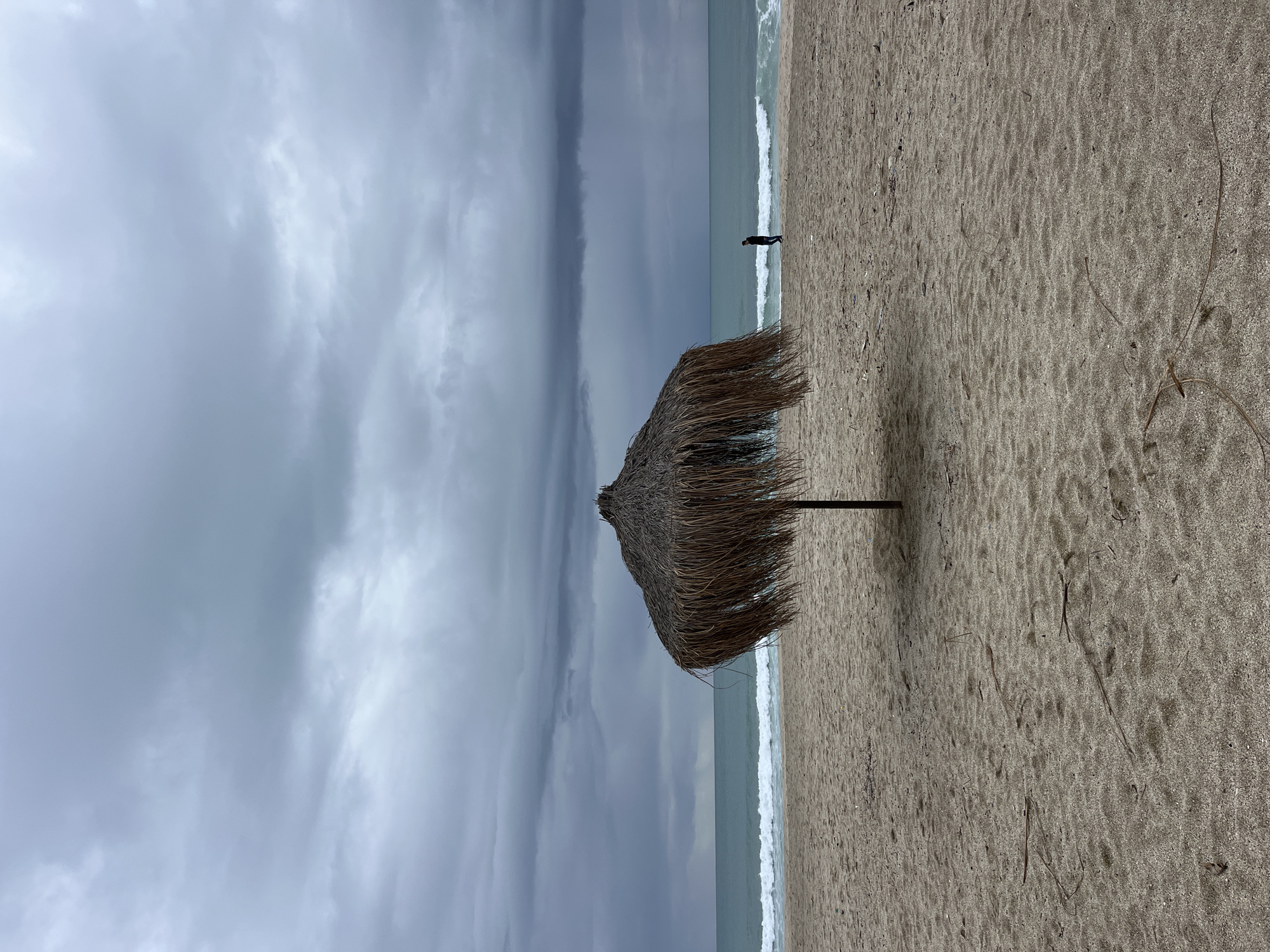 The image captures a beach scene in Alanya, Turkey. A straw beach umbrella stands prominently on the sandy shore. In the distance, a lone individual stands near the water's edge. The sky is overcast with heavy, gray clouds, creating a somber atmosphere. Waves gently break on the beach, marking the boundary between the sand and the sea. The sand appears damp and textured, with scattered debris.
