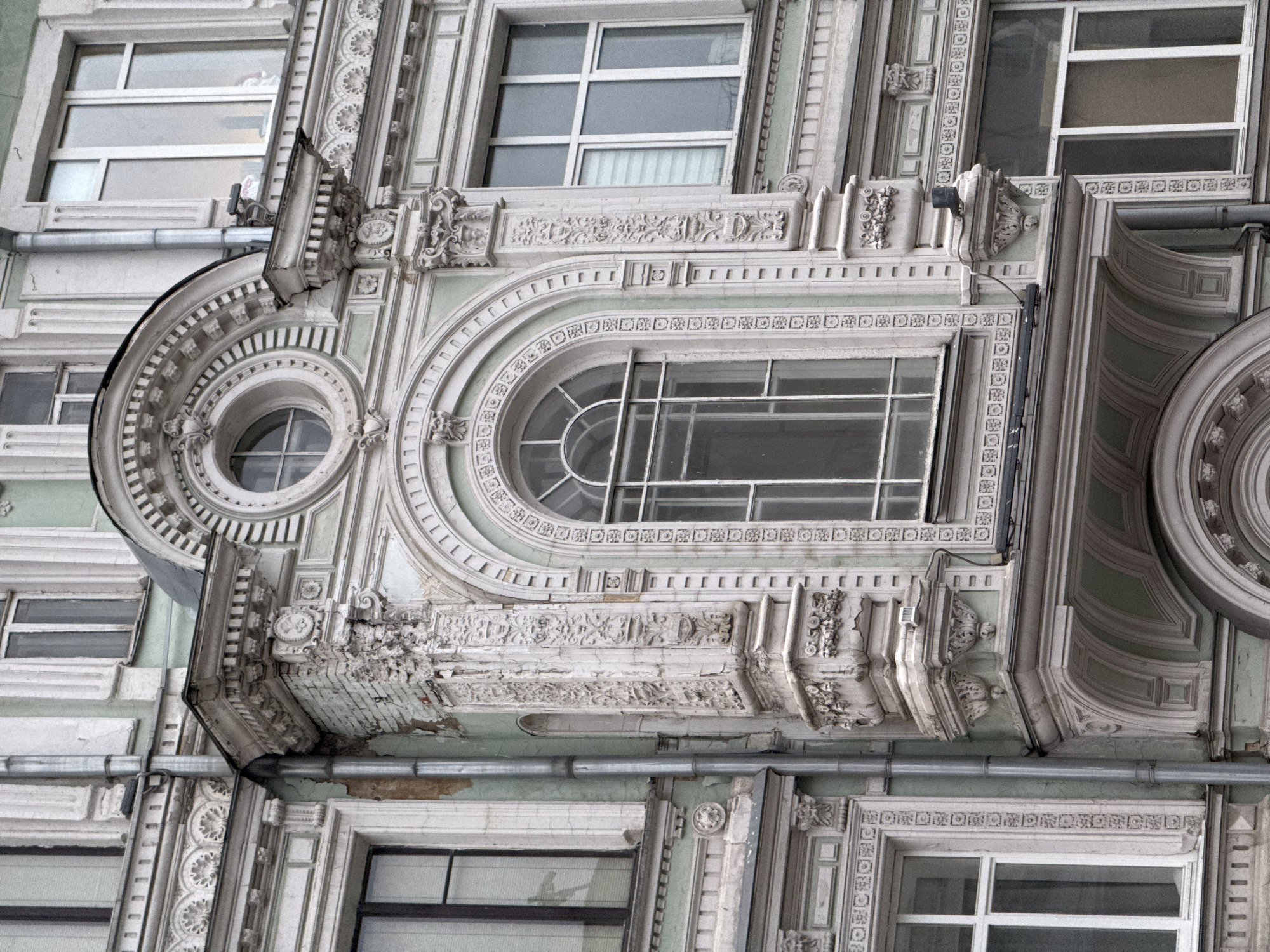The image shows the exterior facade of an ornate building in Moscow, Russia. The architecture is characterized by elaborate decorative details, including moldings, reliefs, and arched windows of varying sizes. The building's color scheme is primarily light, with shades of gray and white. The structure appears aged, with visible signs of weathering and deterioration on the facade. Drainpipes are present along the building's exterior.