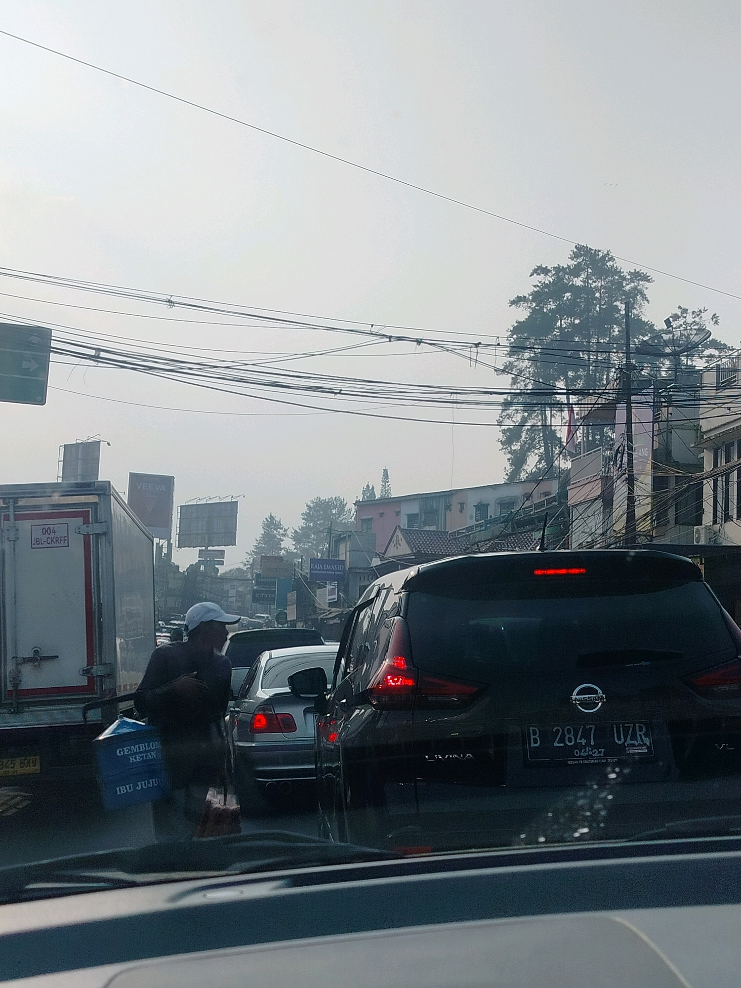 The image captures a congested traffic scene in Cisarua, Indonesia, under a hazy sky. In the foreground, the rear of a dark-colored Nissan Livina is visible, displaying the license plate "B 2847 UZR". To the left, a white truck with the marking "004 JBL-CKRFF" is partially obscured. A person wearing a white cap and a dark shirt stands beside the truck, carrying a blue container with text that reads "GEMBLONG KETAN IBU JUJU". Other vehicles are stalled in traffic, with buildings and trees lining the roadside in the background. Overhead, a dense network of power lines crisscrosses the sky. The overall atmosphere suggests a typical traffic jam in a bustling urban environment.