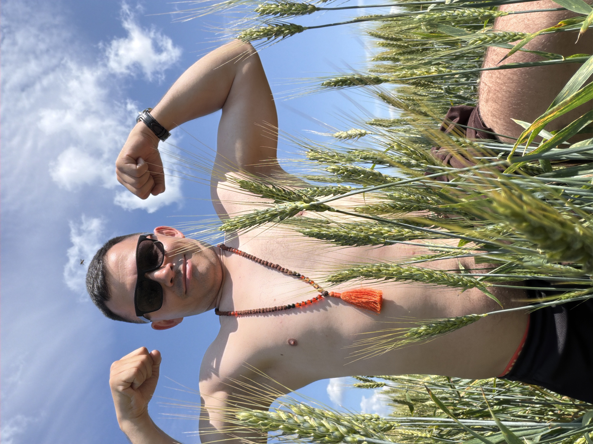 An adult male, with short dark hair and wearing sunglasses and a beaded necklace with an orange tassel, poses in a field of wheat under a bright blue sky with scattered clouds. He is shirtless, displaying his torso and arms, and is flexing his biceps. He is positioned in the foreground, with the wheat stalks obscuring his lower body. The scene is set in Podshivalovo, Russia, suggesting an outdoor, rural location. A small insect is visible in flight in the upper portion of the frame, near the sky.
