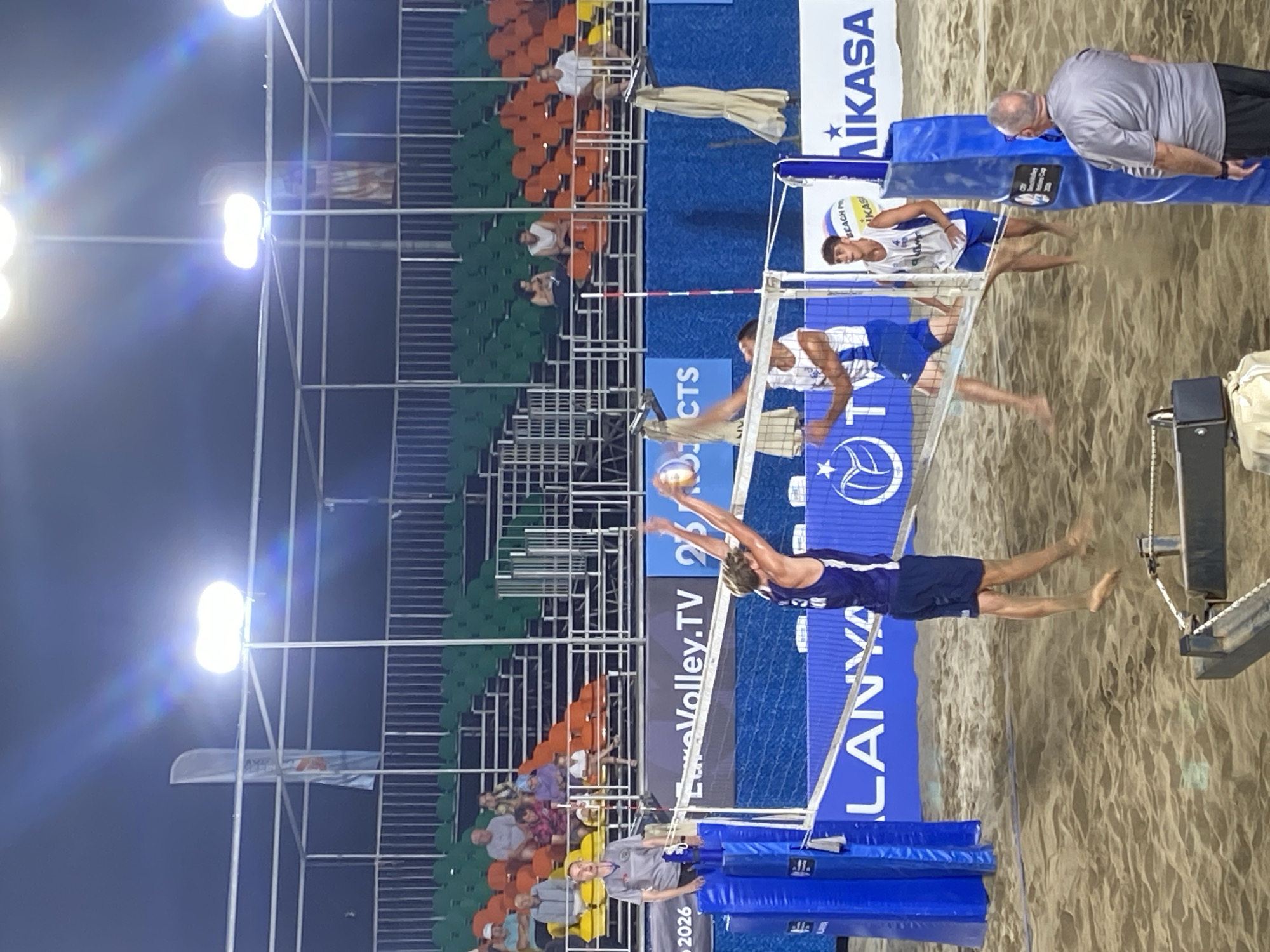 This image captures a nighttime beach volleyball match in Alanya, Turkey, held at an arena under artificial lighting. The court is a sand surface, with a white net stretched across, supported by blue padded poles.
In the foreground on the left, a male player, barefoot and wearing a dark blue/purple sleeveless shirt and matching shorts, is captured mid-air, having just jumped to strike the light-colored volleyball over the net with his left arm. Directly across the net, another male player, barefoot and wearing a white sleeveless shirt with blue accents and blue shorts, is also positioned near the net, with his right arm extended upwards, seemingly preparing to block or react to the ball. A third male player, in a similar white and blue uniform, stands on the sand further to the right, observing the play.
A male referee, dressed in a grey short-sleeved shirt, stands on a blue referee stand at the net pole on the left, intently watching the action. In the bottom right corner, a bald male official, wearing a grey short-sleeved shirt and dark shorts, stands observing the game.
The background features tiered stadium seating filled with spectators, with orange, green, and yellow plastic seats. Above the stands and court, a white metal framework supports multiple bright stadium lights that illuminate the scene, creating visible lens flares against the dark night sky. Banners visible around the court include "EuroVolley.TV," "ALANYA," "MIKASA," and partial text for "CEV Beach Volleyball Nations Cup," confirming the location and type of event. Two light-colored, tall vertical flags and two folded beige umbrellas are also visible in the background.