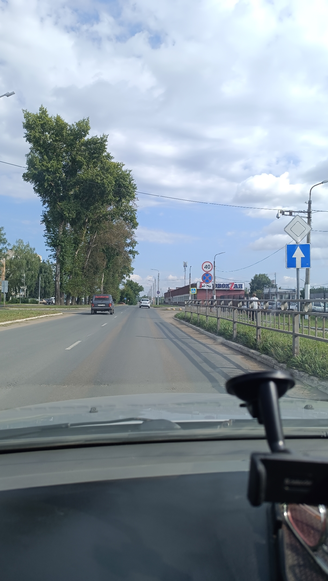 The image depicts a forward view from inside a vehicle traveling on a paved road in Chaikovsky, Russia. The road has multiple lanes and is lined with trees on the left and a metal fence with grass on the right. Several cars are visible on the road, including a dark SUV in front and a white sedan further ahead. Traffic signs are present on the right side of the road, including a speed limit sign indicating 40, a yield sign, and a directional arrow sign. A building with signage is visible in the background on the right. The sky is partly cloudy. A dashcam or similar device is mounted on the dashboard in the foreground.