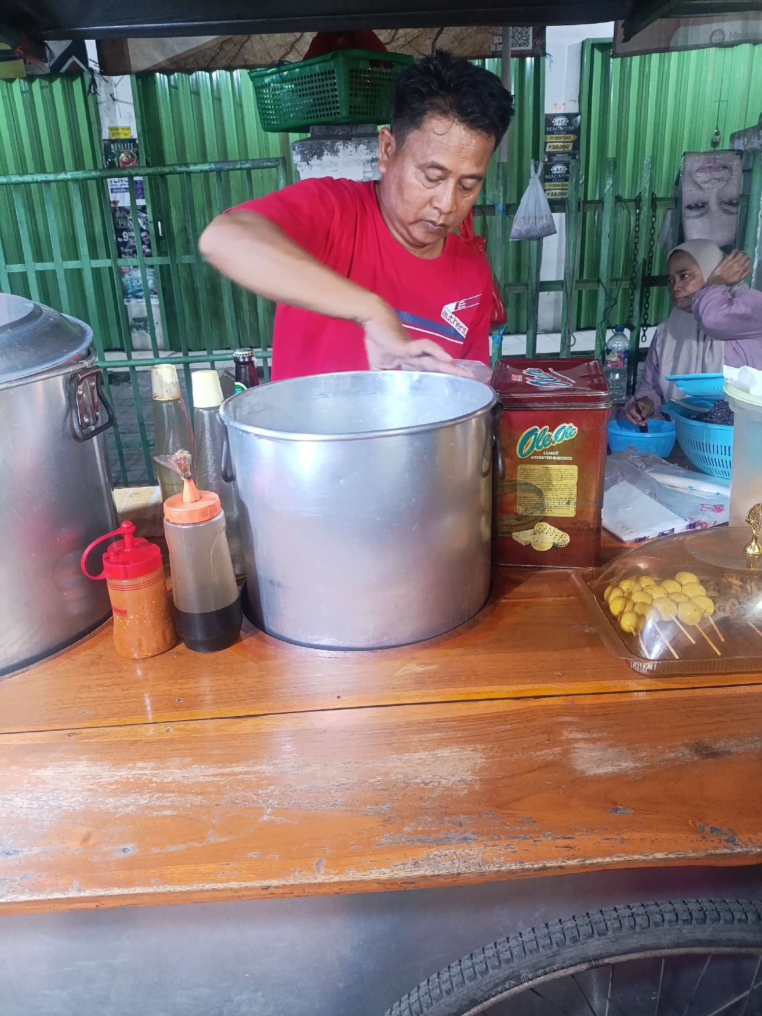 The image depicts a man in a red t-shirt preparing food at a market stall in Jonggol, Indonesia. He is in the process of stirring something in a large aluminum pot. In the background, a woman in a light purple hijab is visible, also at a stall. The stall is made of corrugated metal fencing. Several items are displayed on the counter in front of the man, including a large aluminum pot, smaller sauce bottles, and a tin of biscuits labeled "Ole Ole Family Assorted Biscuits". A clear plastic container with yellow, round food items on skewers is also visible. The counter appears to be made of aged, orange-stained wood. A bicycle wheel is partially visible at the bottom of the frame.