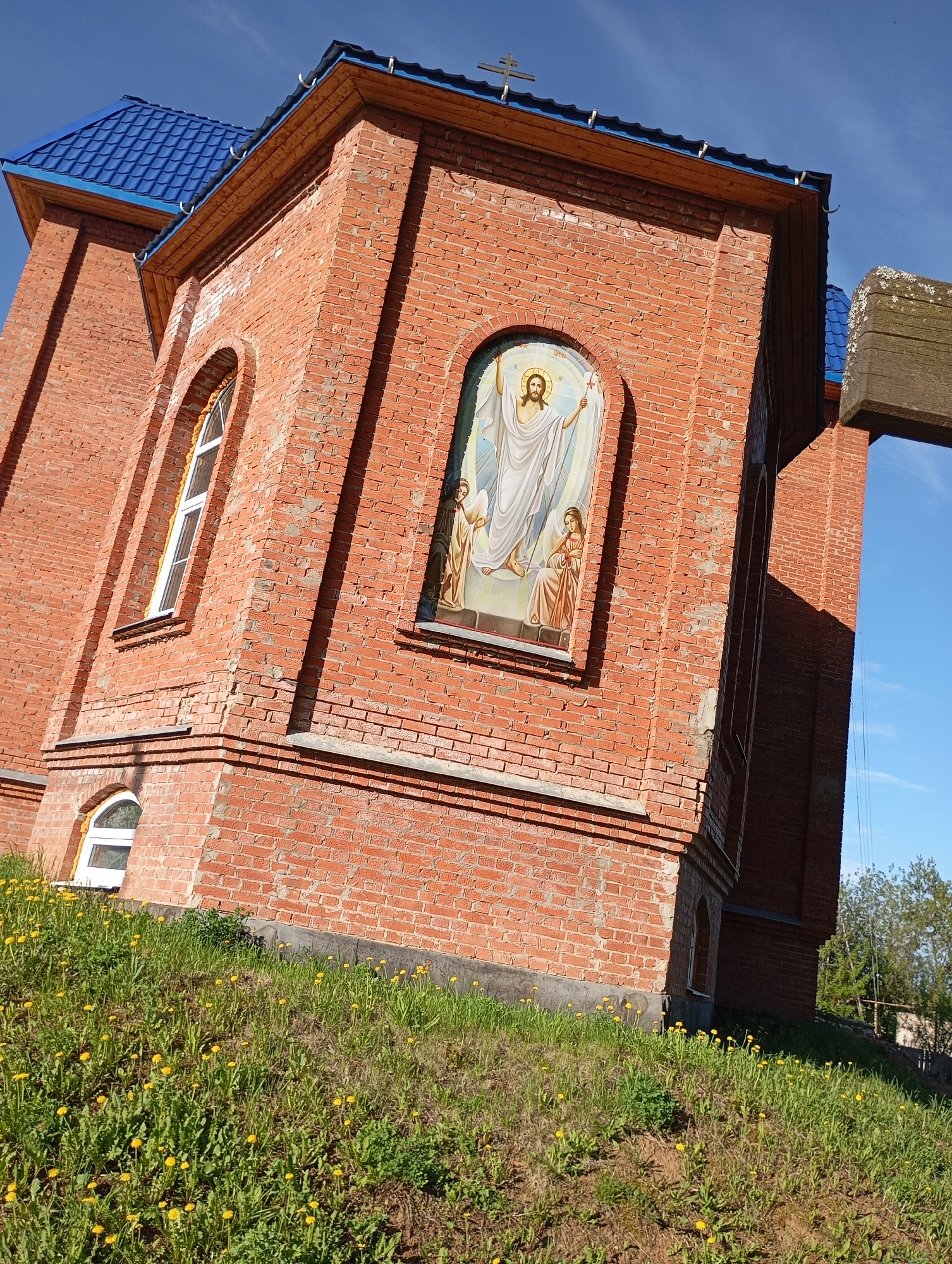 A low-angle shot of a brick building, possibly a church, features a blue roof and arched windows. An icon or religious artwork depicting Jesus and two figures is set into the building's wall. In the foreground, a grassy slope is dotted with yellow flowers. A wooden cross is visible on the right side of the frame against a clear blue sky. The building and lawn are bathed in bright sunlight.