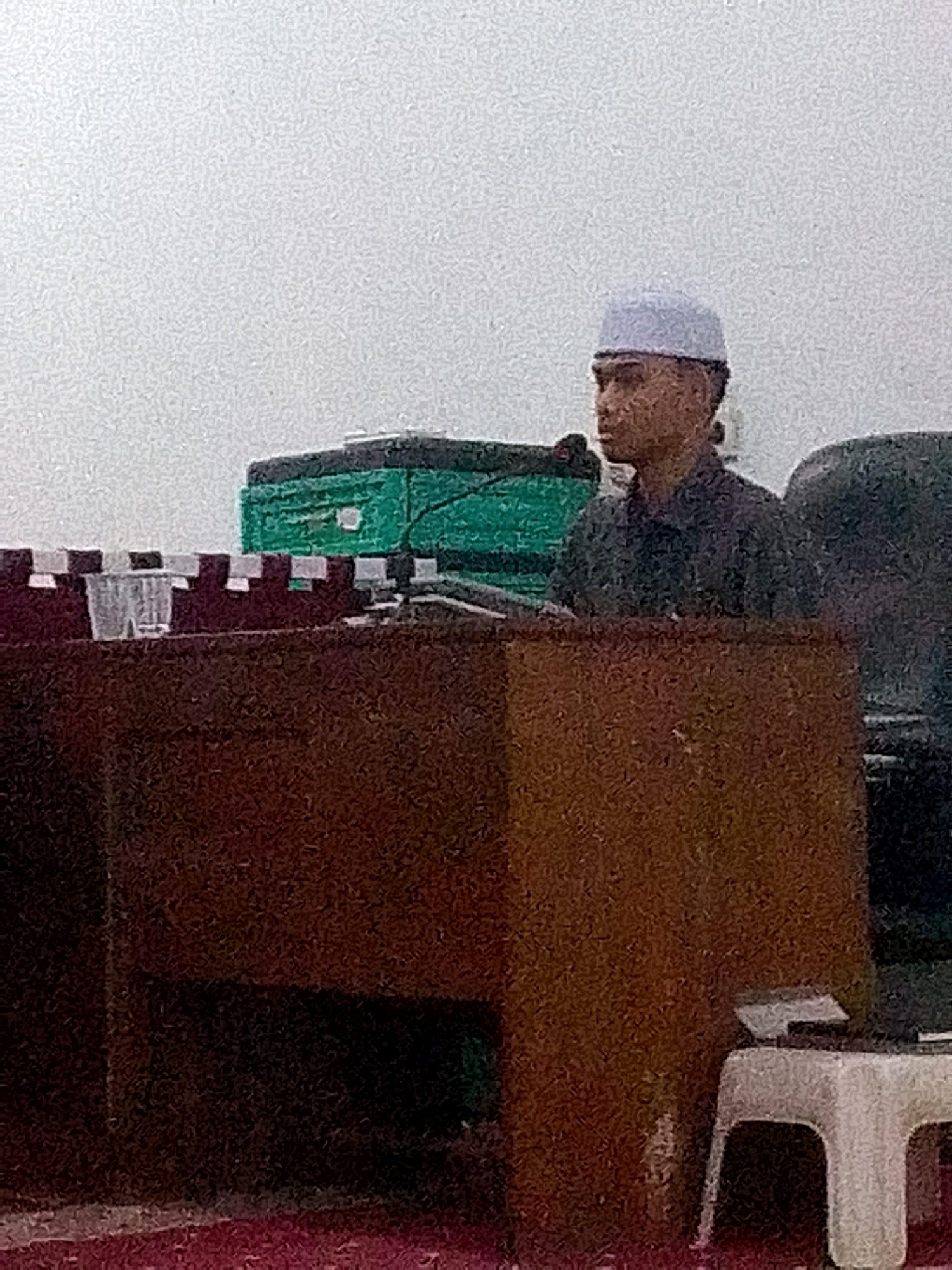 A young man wearing a white skullcap and a dark collared shirt stands behind a wooden podium, facing right. He is positioned in front of a green object that appears to be a banner or backdrop, and there are dark shapes behind him, possibly furniture or other people. To the right of the podium, a white stool with a flat surface is visible. The man is looking to his right, and his left hand is slightly raised. The image has a grainy texture and appears to be taken indoors, possibly in a community hall or religious building. The location is specified as Gunungmasigit, Indonesia.