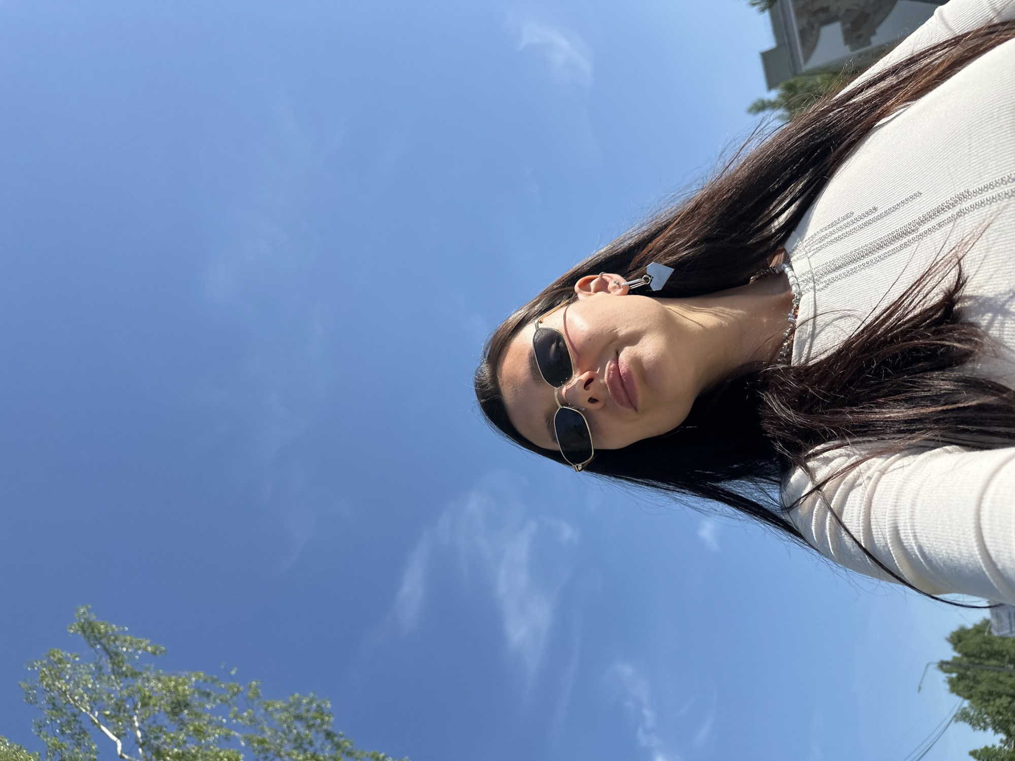 A low-angle, outdoor shot shows a woman from the chest up, wearing sunglasses and a light-colored top, with a clear blue sky in the background. A tree is visible in the lower left corner, and a building is partially visible in the upper right. The woman has long, dark hair. The presence of a building and tree suggests an urban or suburban environment. Given the prompt's indication that the location is Chaikovsky, Russia, it can be inferred the image was taken there, however, without further contextual information it's impossible to determine precisely where in the city this picture was taken.