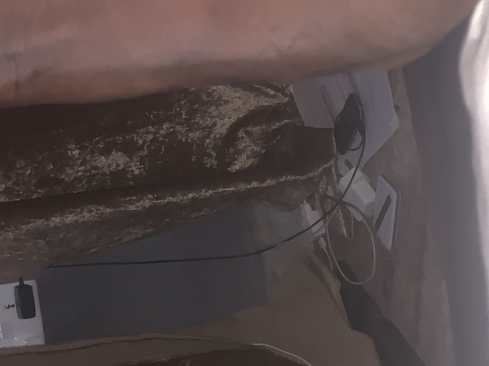The image depicts a close-up view of electronic devices and cables on a textured surface, possibly a floor or table. In the upper left corner, a white electrical outlet with a black adapter is visible. A black cable extends downwards from the adapter, intertwining with other cables and a dark-colored electronic device, likely a smartphone, which is placed on top of a stack of white papers. The background consists of a richly textured, dark fabric, possibly a curtain or upholstery. A blurred human leg and arm are present on the right side of the image, suggesting the presence of a person in the scene. The overall lighting is dim, with a slight warm cast. The location is Jalingo, Nigeria.