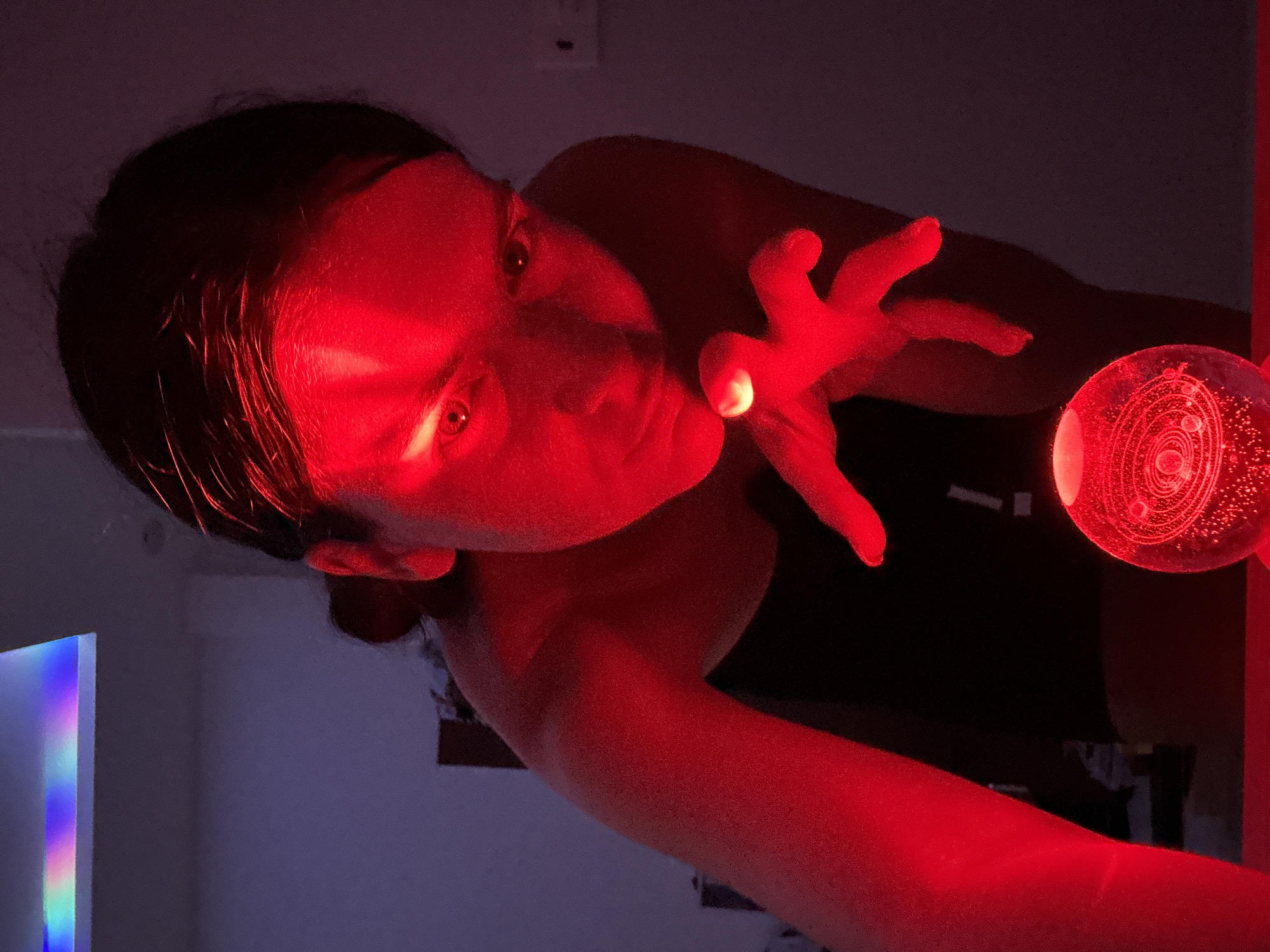 A young woman with dark hair is illuminated by red light, leaning forward with her face close to a glass orb that displays a solar system model. Her right hand is extended, fingers splayed, with a bright light emanating from her fingertip, creating a halo effect. The orb sits on a red surface, and the background is dark and out of focus, with subtle purple hues and a strip of rainbow light on the ceiling. The scene appears to be indoors, possibly in a bedroom or a dimly lit room. There is no indication of the city of Mũi Né, Vietnam.