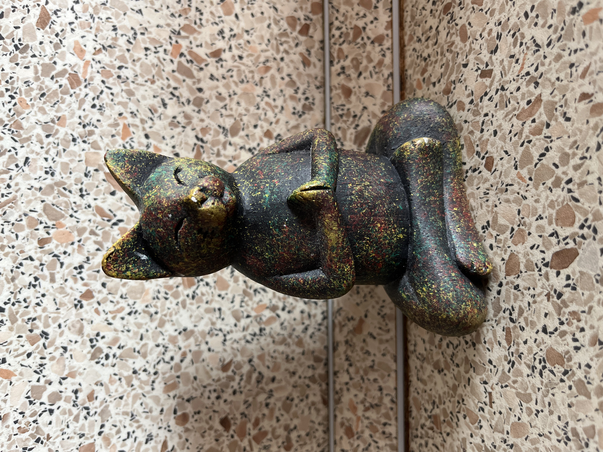 A cat figurine is positioned in a meditative pose on a speckled surface. The figurine is dark with colorful speckles of red, yellow, and green. The cat has its eyes closed and its front paws pressed together in front of its chest. The background is a wall with a similar speckled pattern. The scene does not contain any information about Saratov, Russia, or any other location.