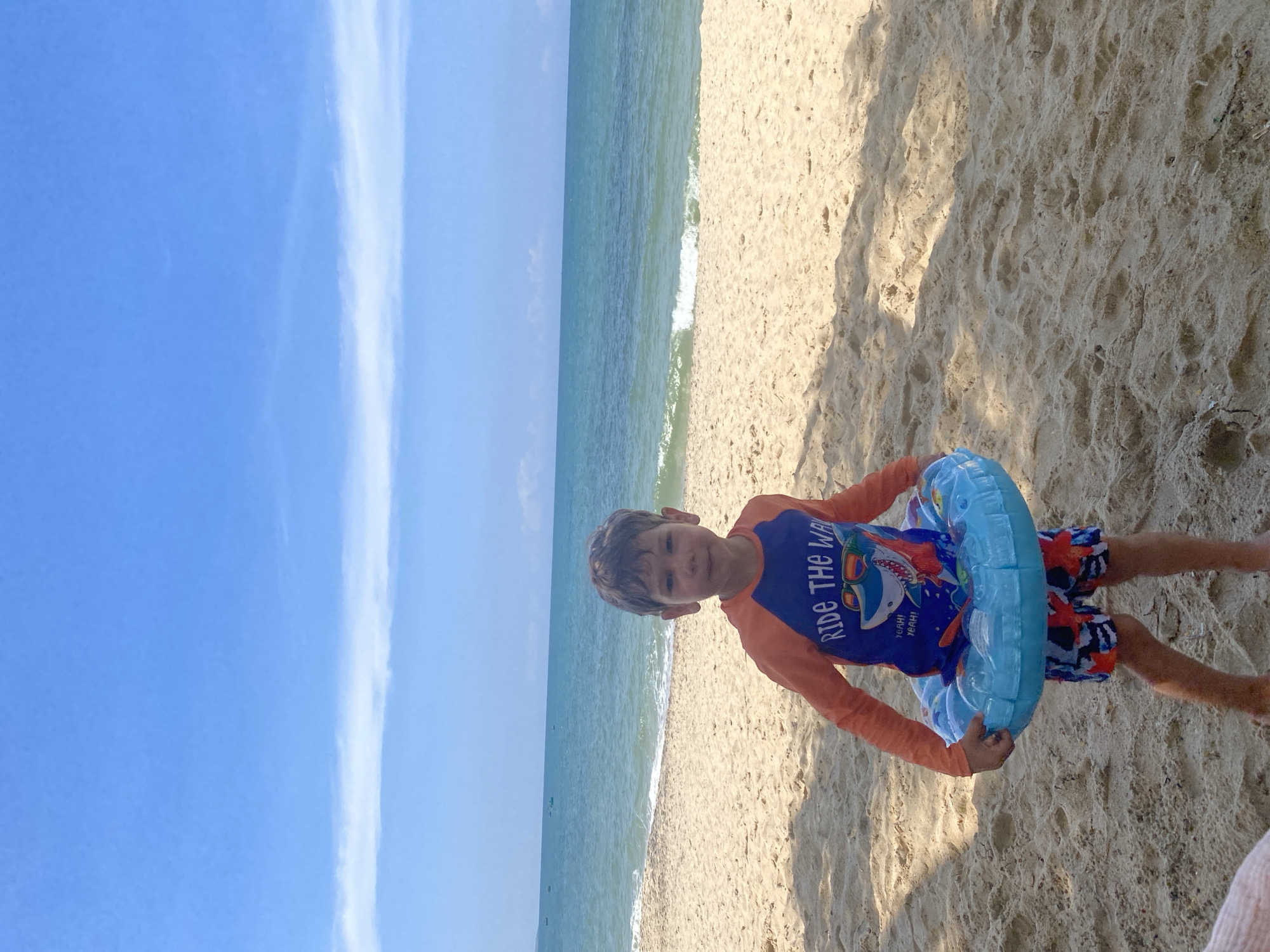 An outdoor, eye-level shot captures a young male child standing on a sandy beach in Phu Thuy Ward, Vietnam, on a bright, sunny day. The child, estimated to be between five and eight years old, has short brown hair and looks directly at the camera with a neutral expression. He is wearing an orange long-sleeved rash guard with a blue central panel featuring a graphic of a shark wearing sunglasses and the text "RIDE THE WAVE," along with patterned swim shorts in red, blue, and white. He holds a light blue inflatable swim ring around his lower torso with both hands. The foreground consists of light brown sand with visible footprints and textural variations, marked by shadows in the lower left and right. Behind the child, the turquoise ocean extends horizontally, with small waves breaking near the shore. Several small, dark-colored boats are visible in the far distance on the water. The upper portion of the image is dominated by a clear, vibrant blue sky with a few faint, elongated white clouds forming a horizontal band.