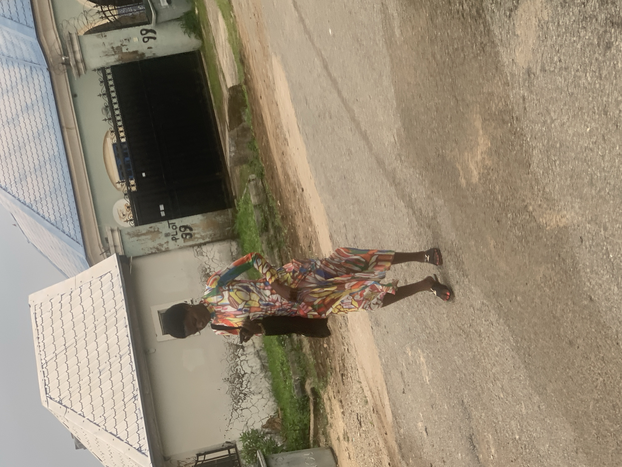 An individual, appearing to be a young woman with dark hair and skin, walks on a paved road, looking down at a mobile phone in her left hand. She is wearing a long-sleeved, multi-colored dress with a high neckline, featuring a vibrant abstract pattern of red, yellow, blue, and black. A dark tote bag hangs from her left shoulder. She is wearing open-toed sandals with dark straps.
The background shows a street scene. To the left, a light-colored building with a white, corrugated metal roof is visible. Further back, to the right, is a taller building with a light blue, sloped roof. A black metal gate with ornate detailing is in front of this building, flanked by pillars. The number "99" is spray-painted in black on one of the pillars and on the wall to the left of the gate, with "PLOT 99" also visible on the wall. The area appears to be a residential street, possibly in the city of Abuja, Nigeria, given the architectural style and the presence of street numbering. The ground is a mix of asphalt and dirt. The sky is overcast.