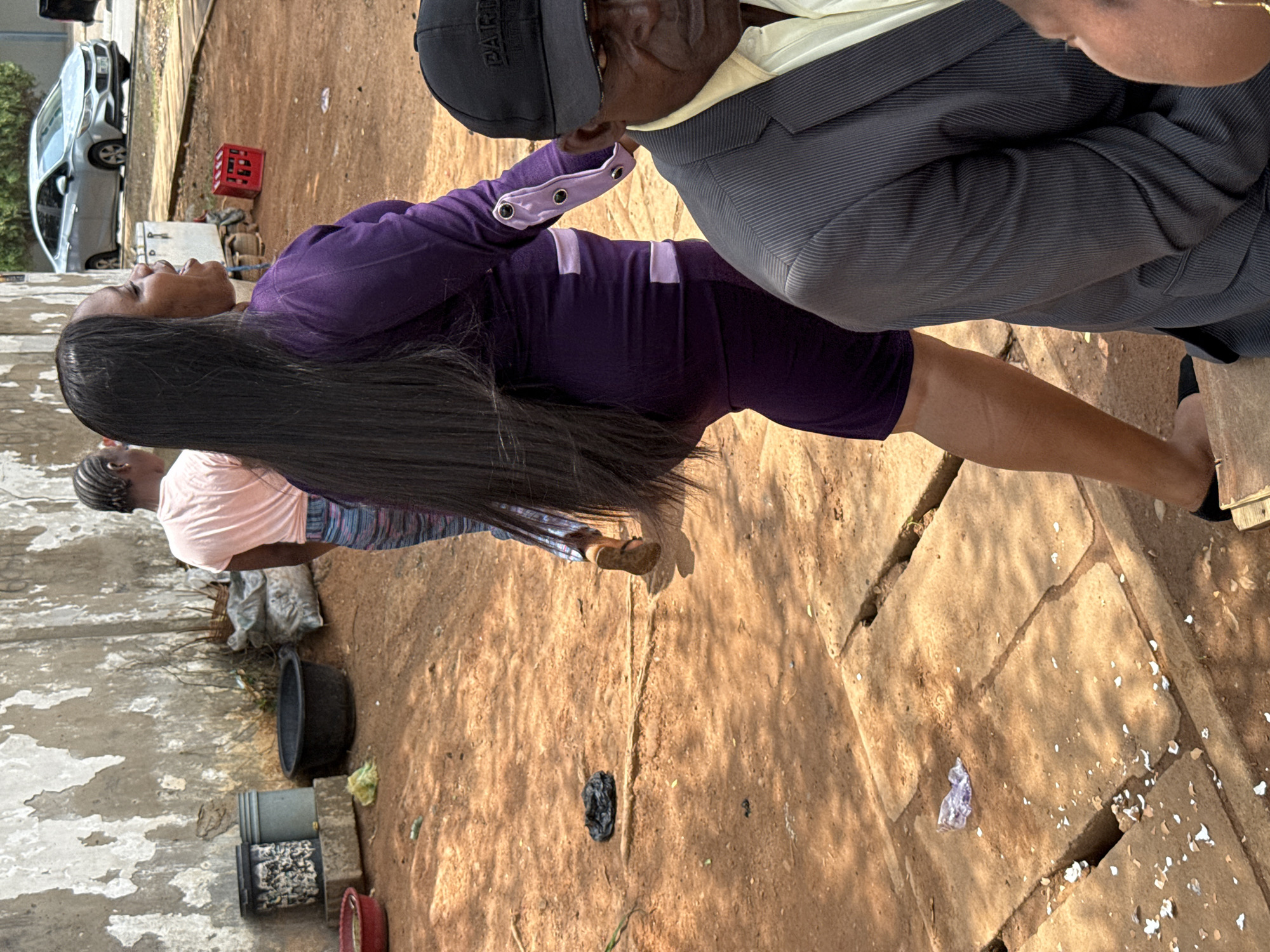The image shows three people in an outdoor setting. A woman in a purple dress with long black hair is standing beside a man in a grey suit and a black cap. A second woman in a pink shirt stands behind the first woman. The ground is dirt and concrete. In the background, there are containers, a red crate, and a silver car. The location is possibly in Abuja, Nigeria.