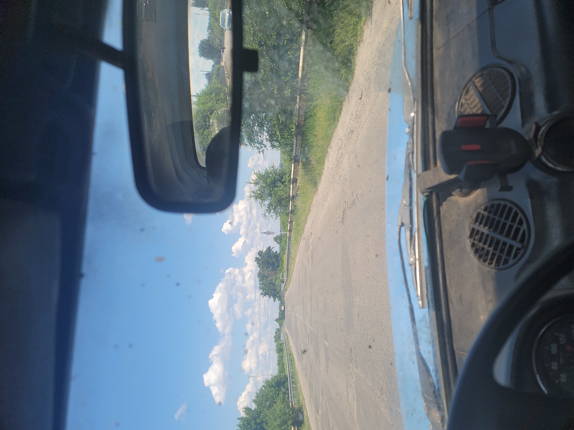 The image is taken from the driver's perspective inside a car on a rural road outside the city of Pereshchepyne, Ukraine. The road is paved but appears worn with cracks and potholes. Guardrails line both sides of the road, and lush green trees and vegetation are visible. The sky is blue with scattered white clouds. In the distance, a white car is approaching and a bridge is visible. The interior of the car shows a portion of the dashboard with a phone mount, a speaker grille, and a steering wheel. The windshield is dusty and streaked with some dark spots, possibly dirt or debris. The rear-view mirror reflects the road ahead, showing more trees and a few other vehicles. The scene suggests a typical daytime drive through a countryside area near Pereshchepyne.