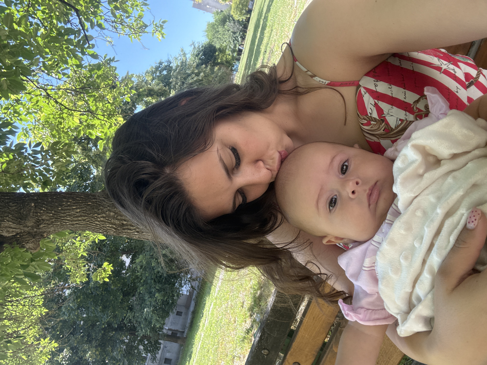 A woman with long, dark hair is kissing a baby on the forehead. They are sitting on a wooden park bench under a large tree. The woman is wearing a red and white striped top with a patterned design. The baby is wrapped in a light-colored blanket and wearing a pink garment. The background shows a grassy park area with more trees and a clear blue sky. The presence of park benches and mature trees suggests a park setting within the city.