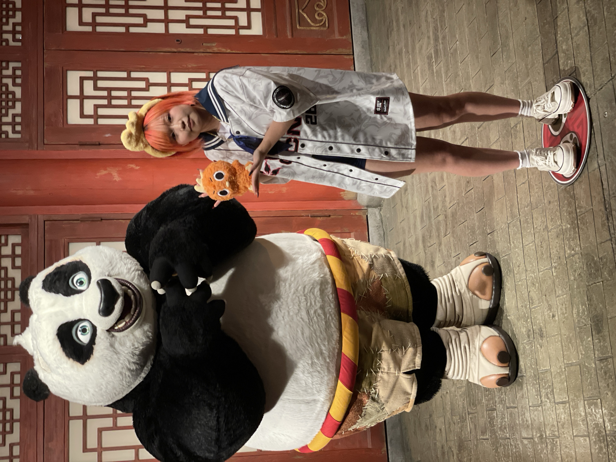 A full shot depicts a person and a panda character in Tongzhou District, China. The individual, standing on a round platform marked with Chinese characters, has orange hair, wears a white baseball jersey-style shirt, and holds a small orange plush toy. Beside them stands a large panda character from "Kung Fu Panda," complete with a hula hoop-like belt and oversized martial arts shoes. The setting features traditional Chinese red doors with intricate wooden patterns in the background and stone brick flooring. The lighting appears to be artificial, suggesting an evening or indoor scene. The person interacts with the panda character by holding the plush toy near it.