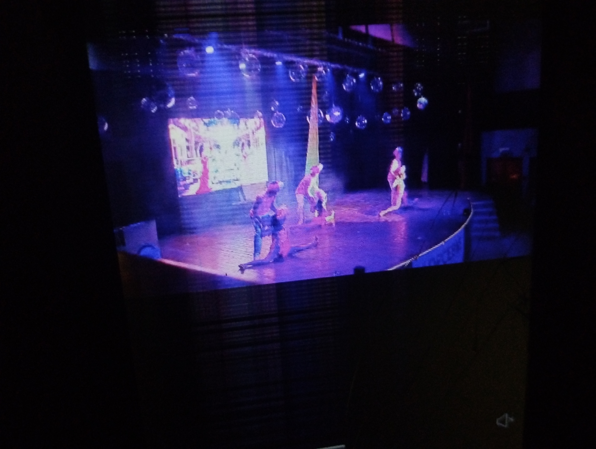 The image shows a stage performance with three dancers illuminated by blue and purple lighting. There are disco balls hanging above the stage, and a backdrop with a picture. One of the performers is interacting with a suspended piece of fabric. The scene appears to be recorded from a screen or monitor, as evidenced by the visible lines and player control at the bottom right. The location, Porlamar, Venezuela, is not discernible from the image itself.